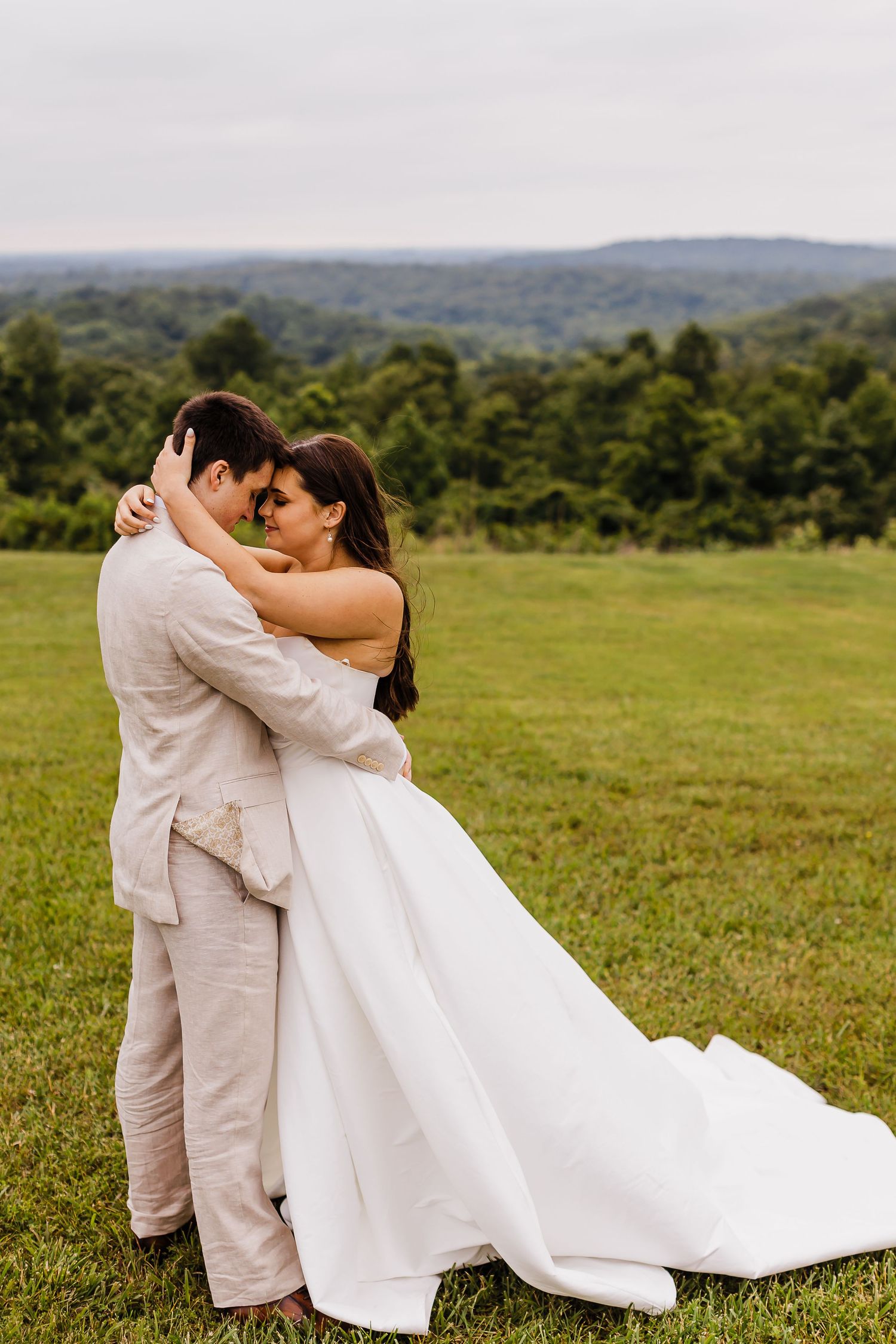 A couple embraces in a romantic moment on a grassy hilltop overlooking a scenic forested landscape.
