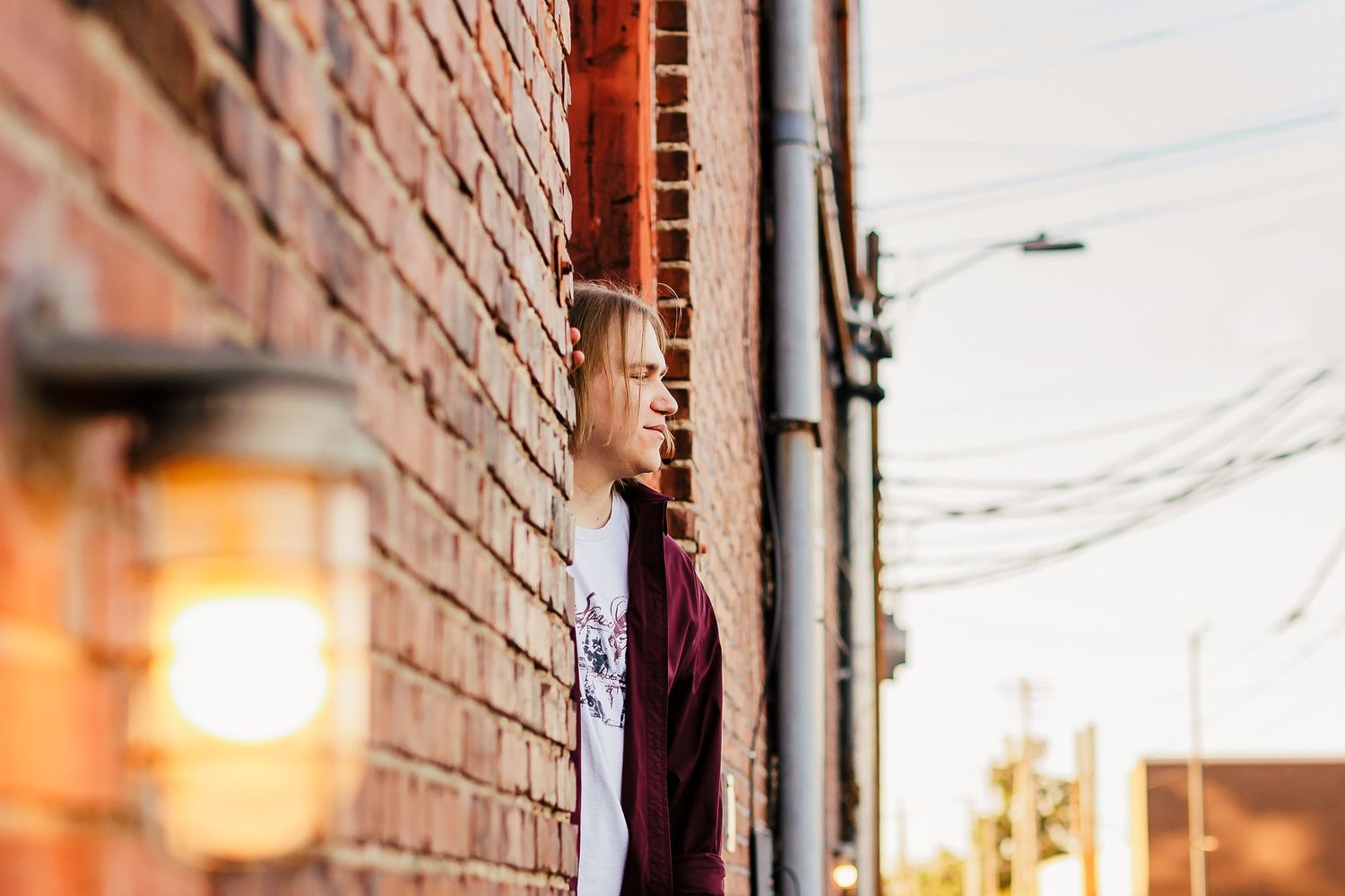 A person peeks out from behind a brick wall at sunset with a glowing street lamp.