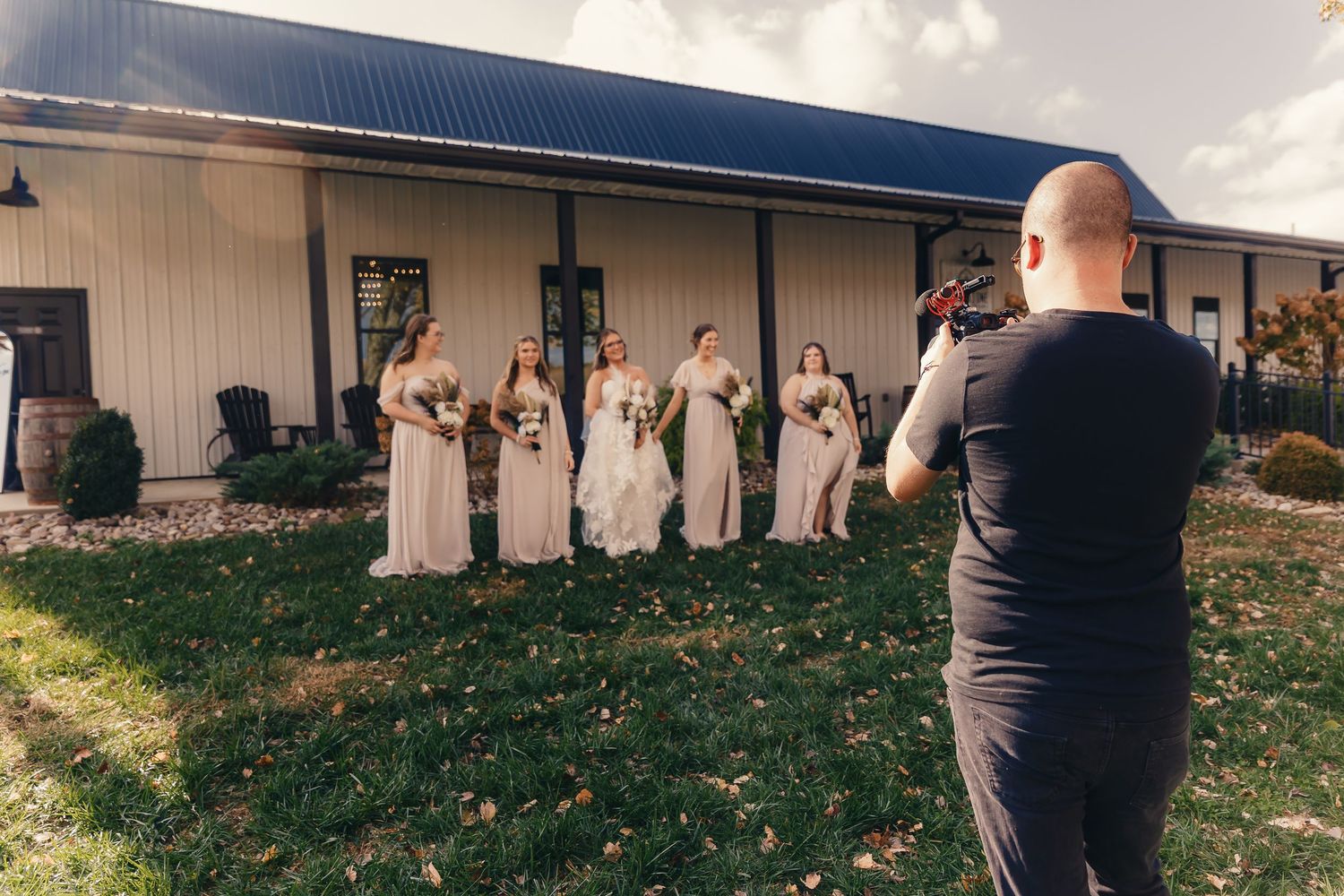A photographer in black captures a wedding party lineup outside a rustic building on a grassy lawn.