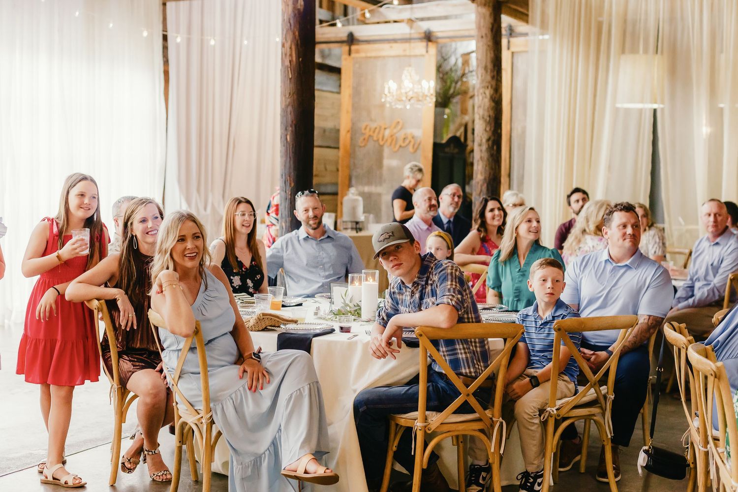 Wedding guests laugh and enjoy themselves during a reception at a bright, elegant venue with wooden chairs and white drapes.