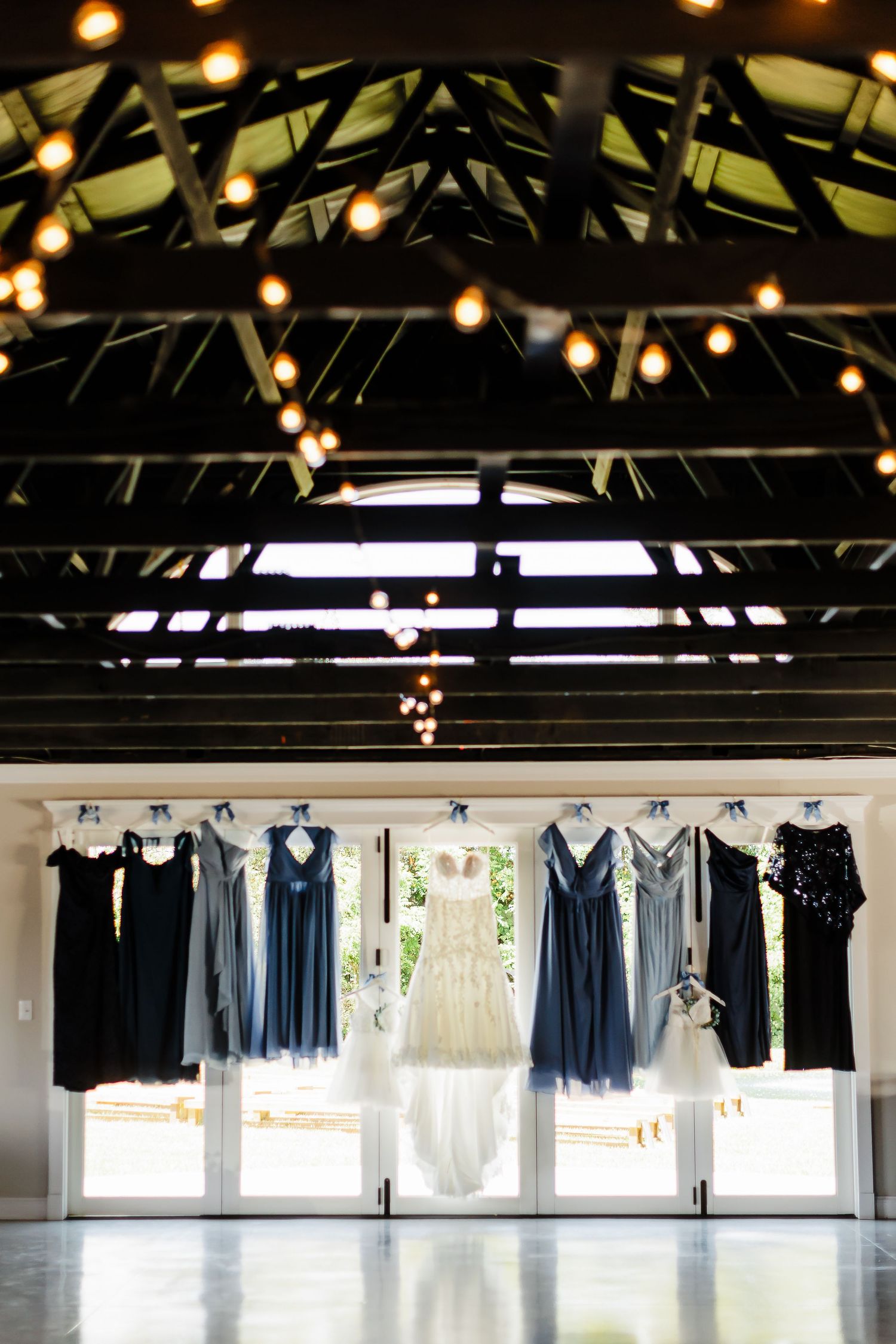 Wedding party dresses in shades of blue and gray hang against a window with string lights above.