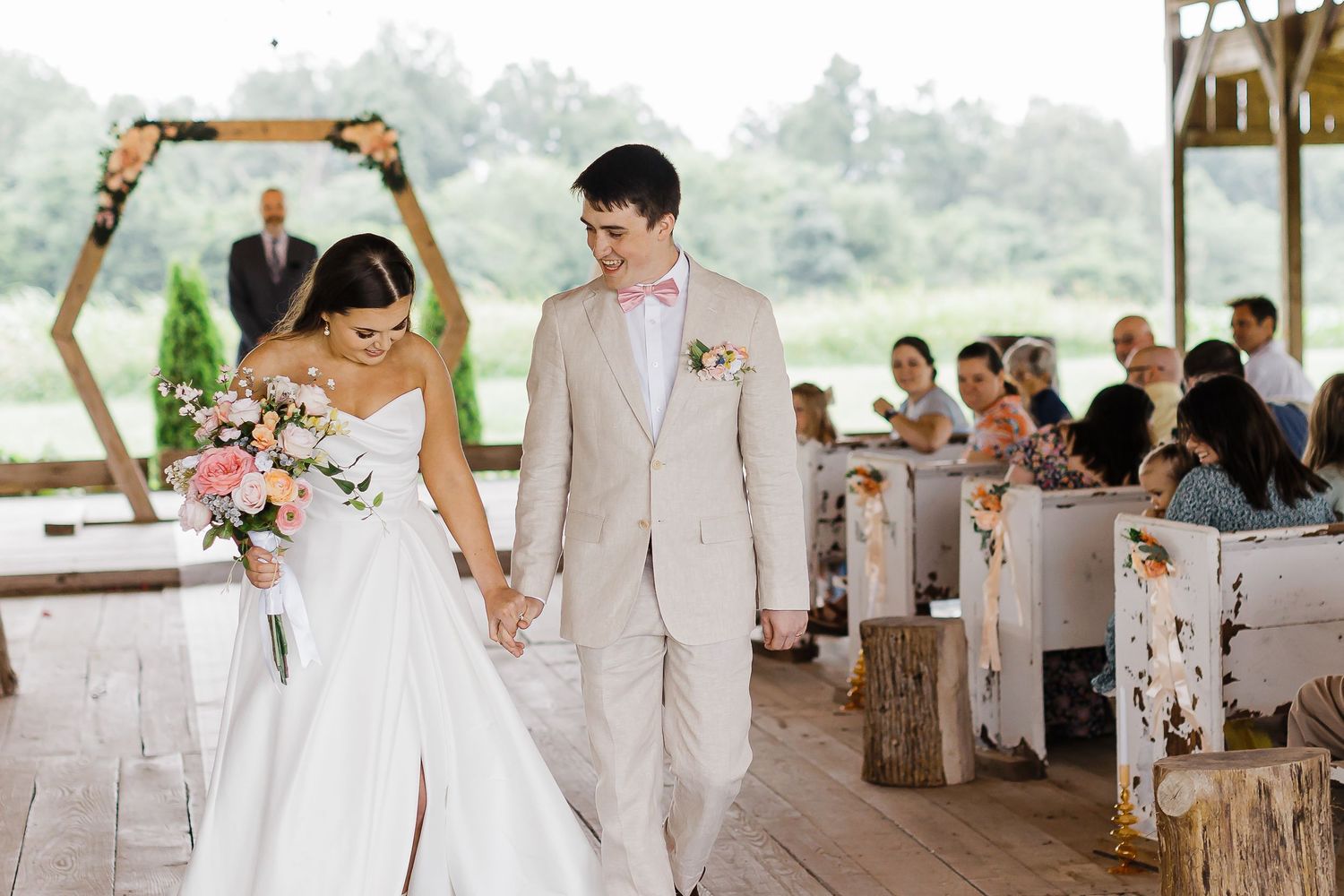 A wedding ceremony takes place under a wooden arch with guests seated on rustic benches.