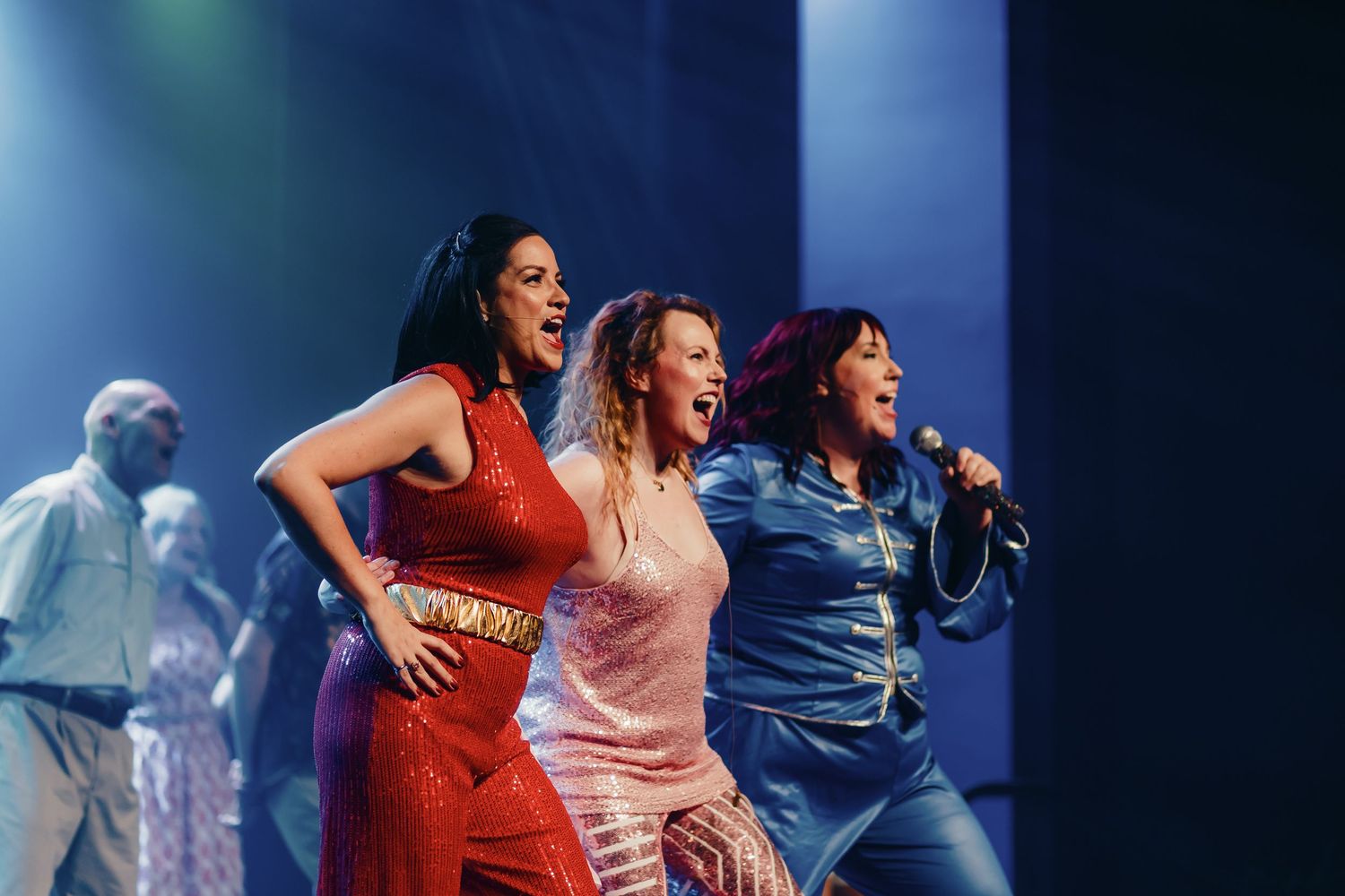 Three women performers sing on stage in colorful outfits under blue stage lighting during a live musical show.