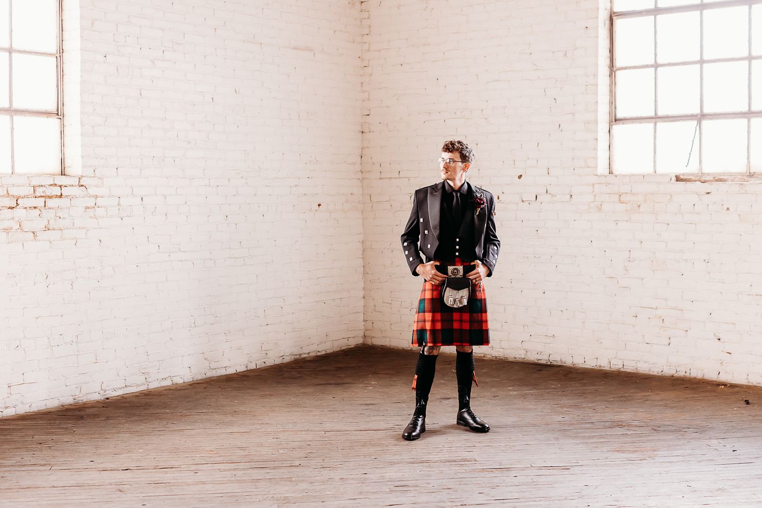 Man in traditional Scottish kilt standing in empty white brick room.