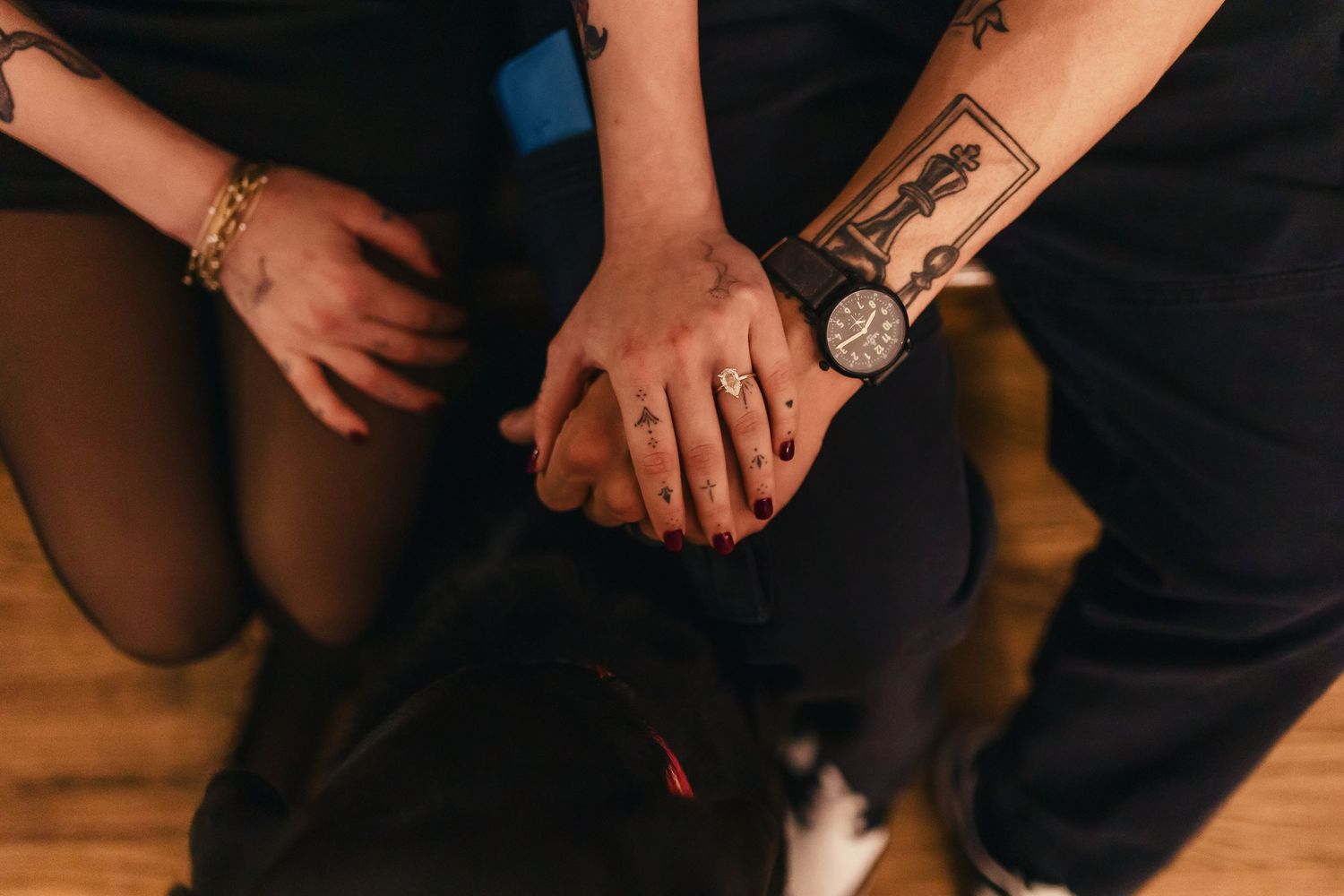 Tattooed hands with a wristwatch holding each other on dark clothing against wooden flooring.