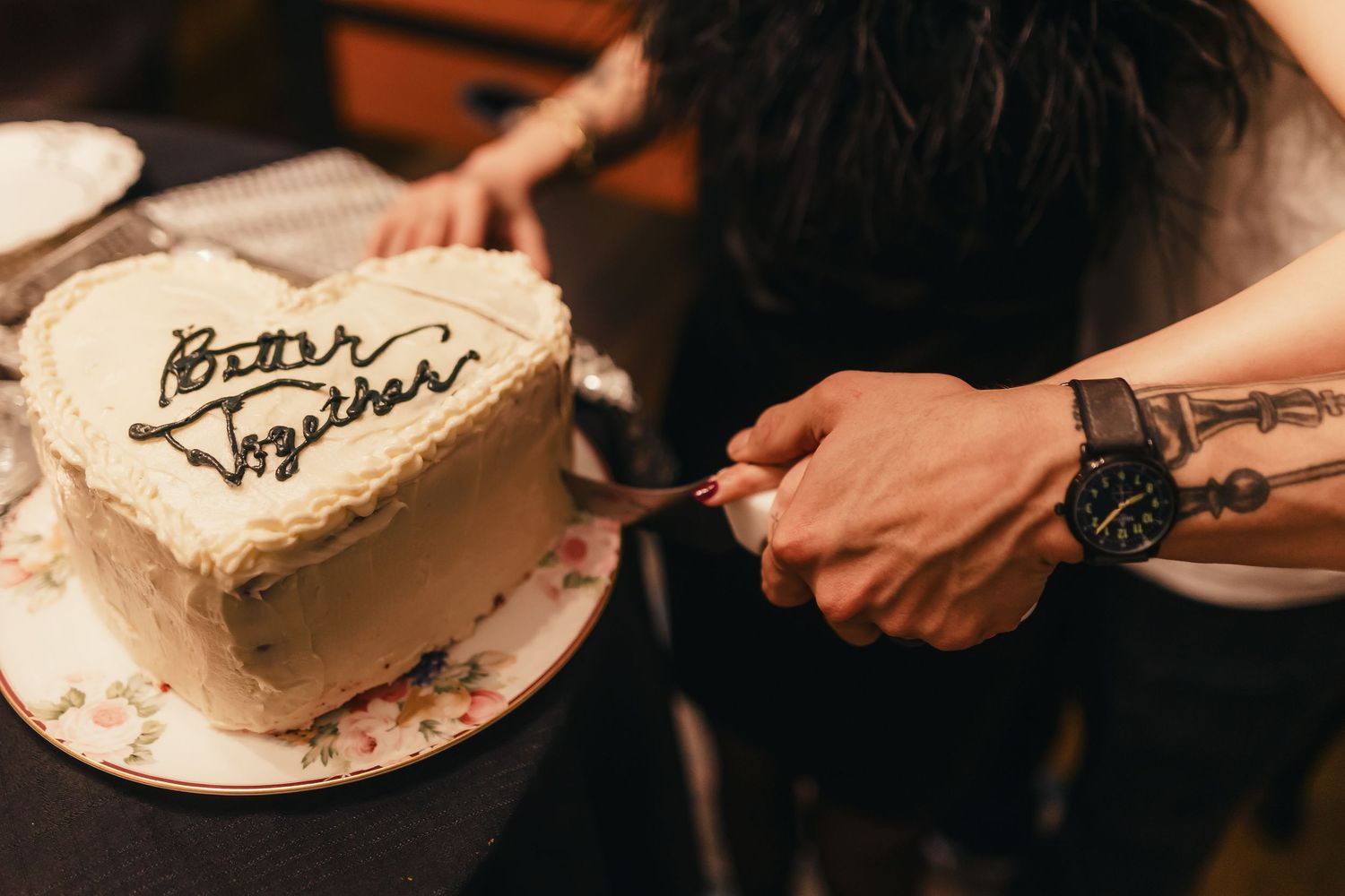 Someone with a tattooed arm cuts into a heart-shaped white cake with decorative writing on top.