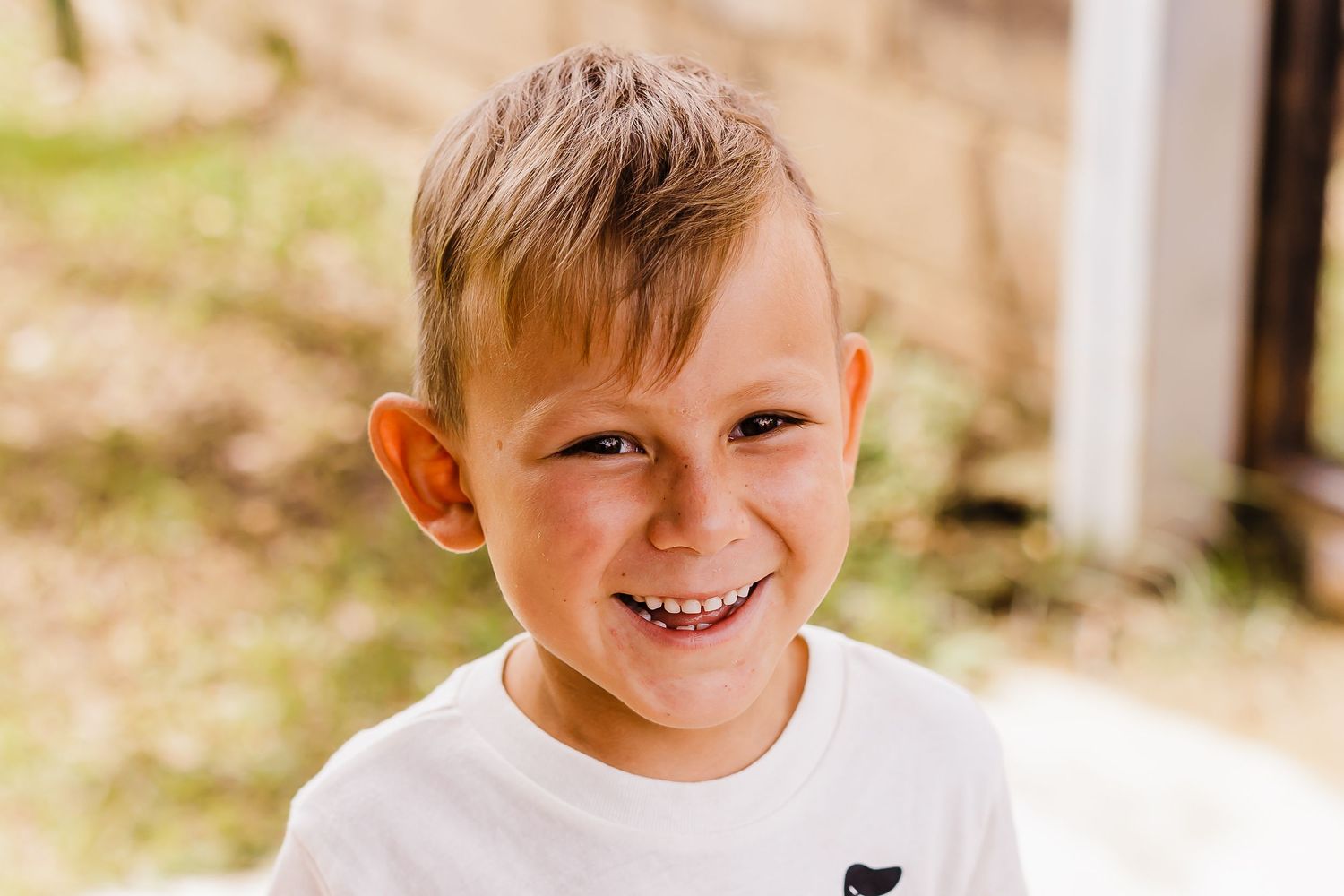Young boy in white t-shirt smiling brightly in outdoor setting.