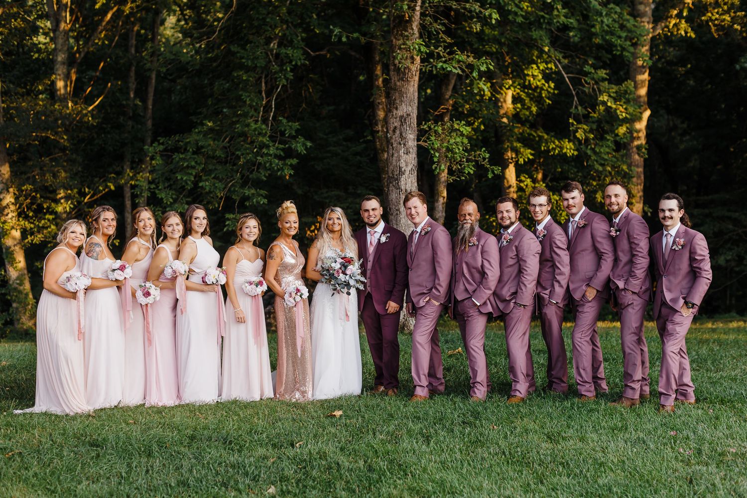 Wedding party poses outdoors with bridesmaids in pink dresses and groomsmen in mauve suits on a grassy lawn.