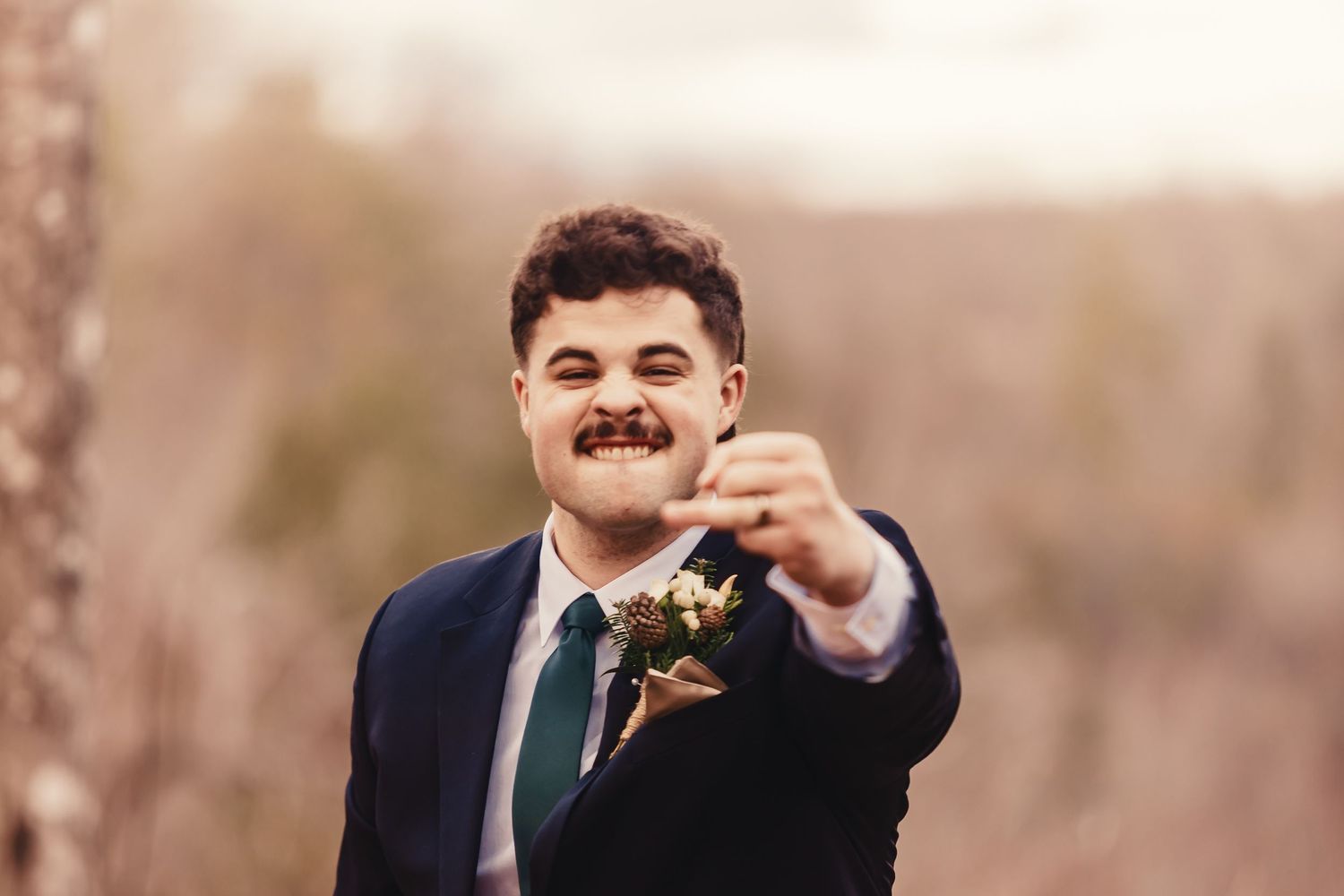 Smiling groom in navy suit and green tie holding a boutonniere outdoors at a wedding celebration.