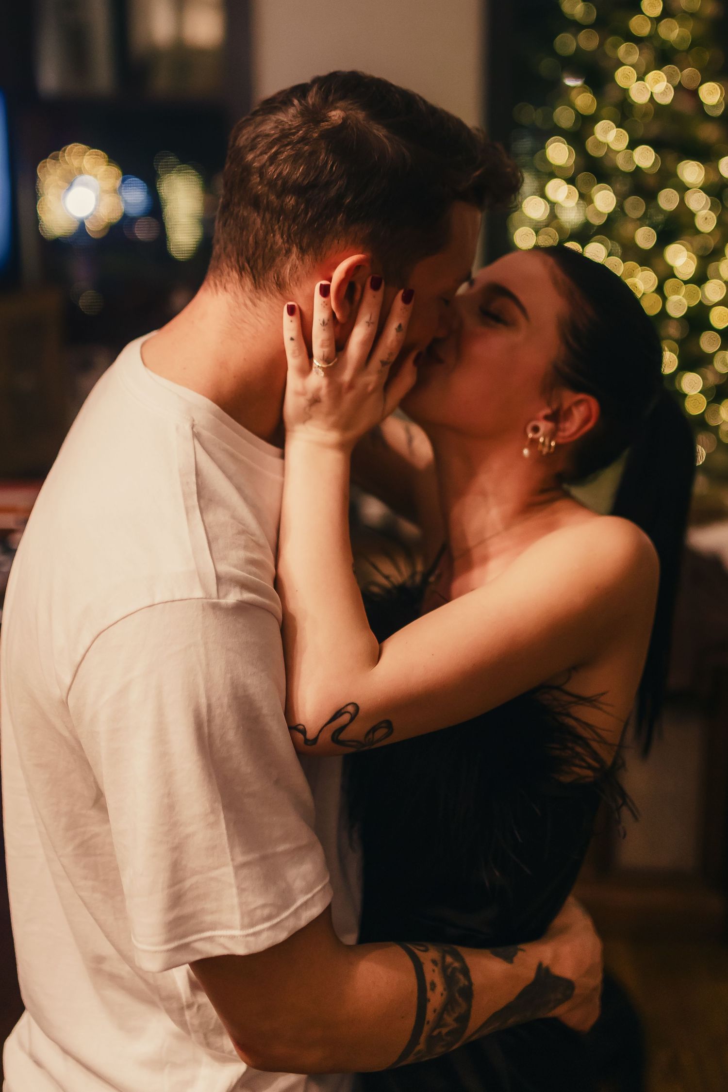 A romantic couple sharing an intimate kiss in front of twinkling Christmas lights in a dimly lit setting.