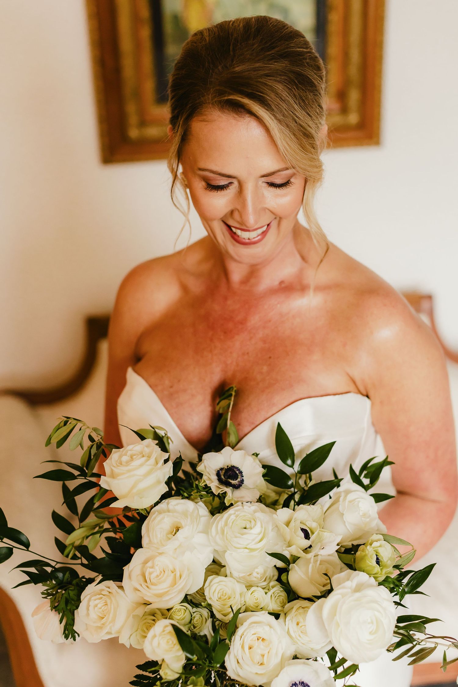 Bride holding elegant white rose bouquet with greenery while smiling in natural light.
