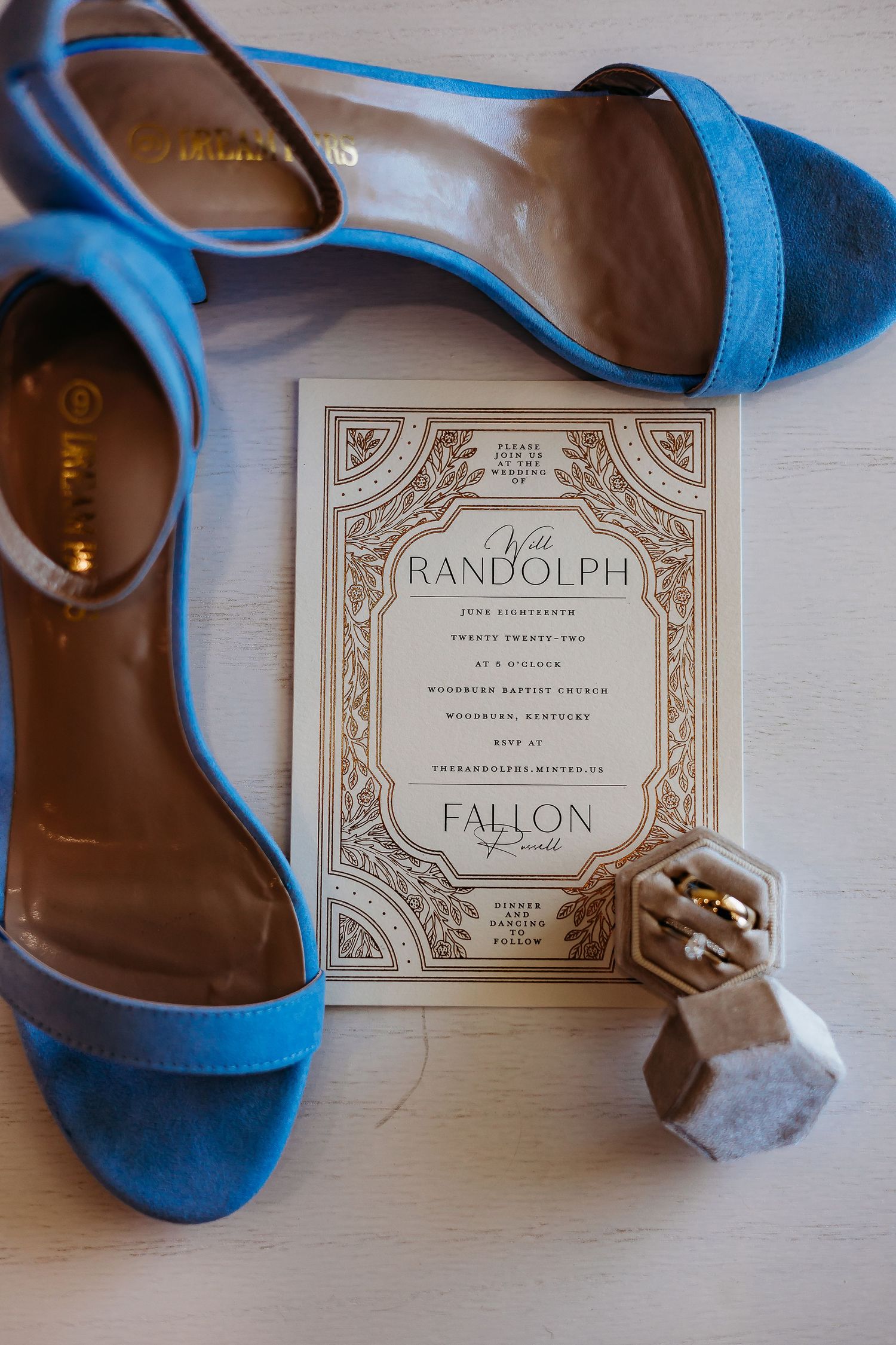Elegant blue suede sandals next to an ornate wedding invitation and small decorative object on white surface.