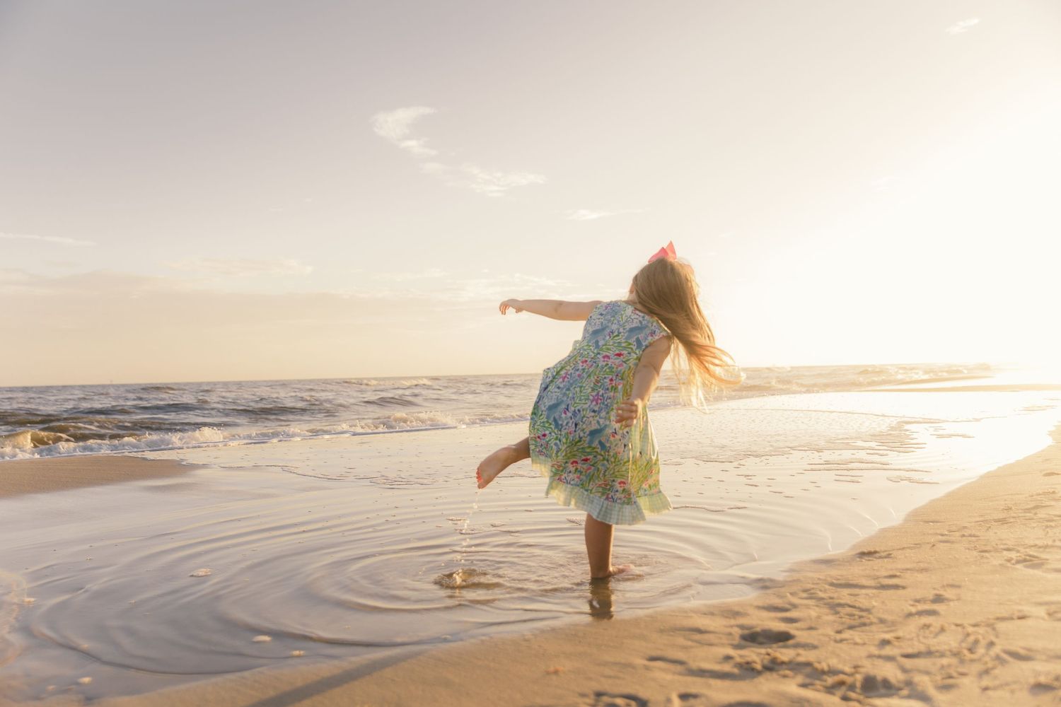 Child twirls in floral dress on beach during golden hour with family in background.