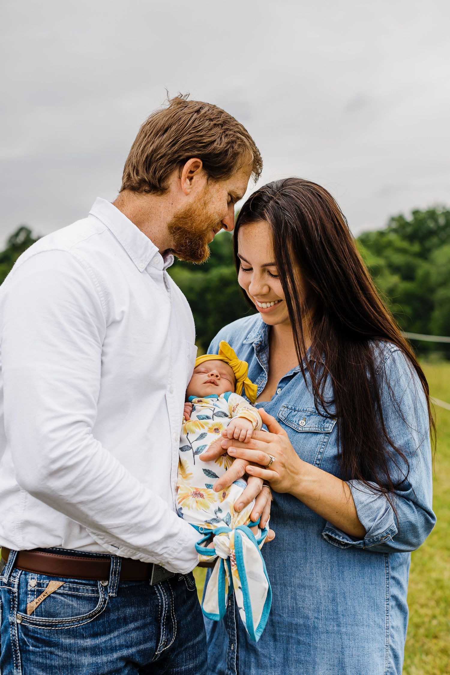 Happy couple smiling at their newborn baby wrapped in a colorful swaddle outdoors on a green field.