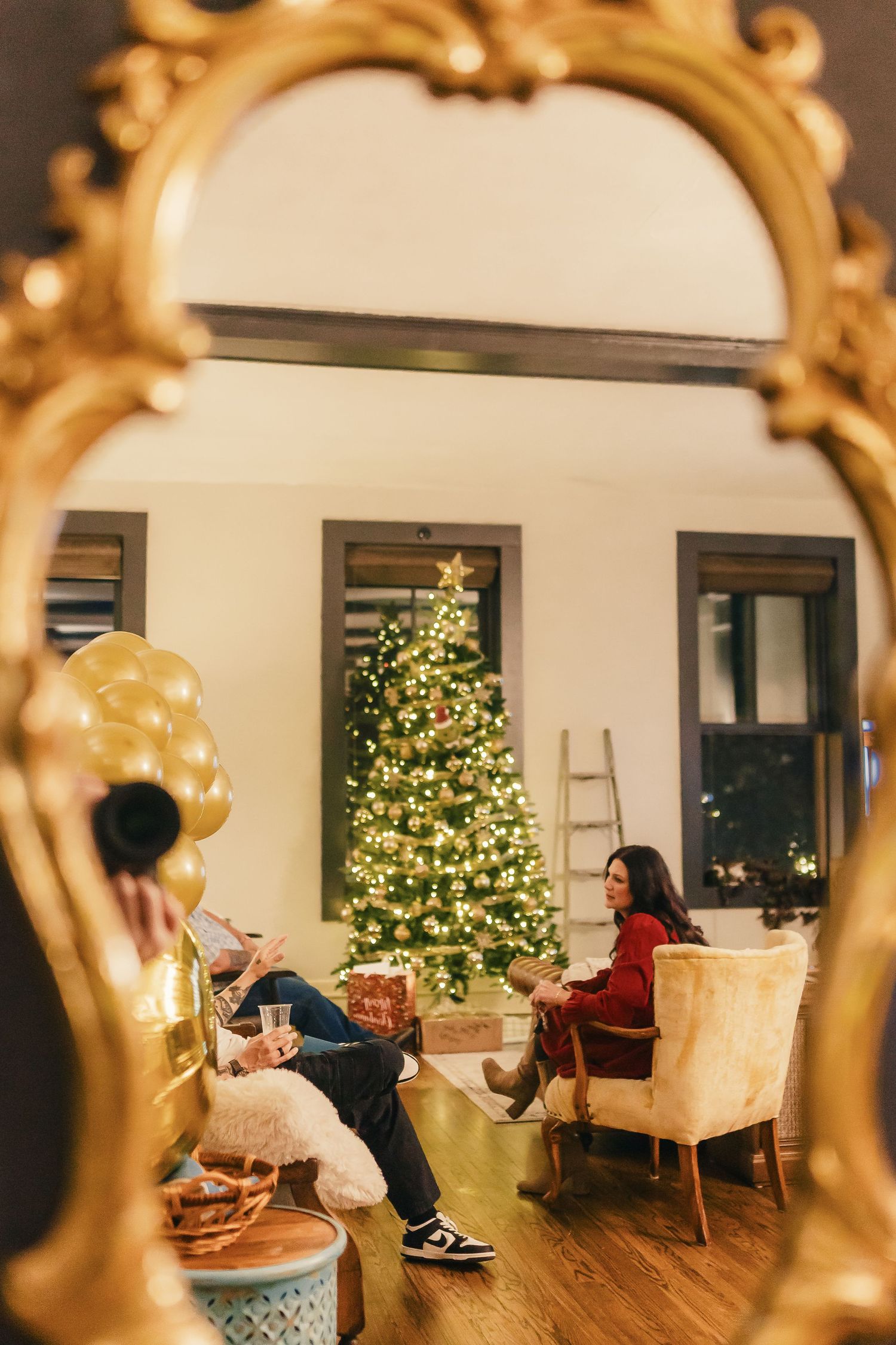A cozy holiday scene viewed through an ornate gold mirror frame showing a lit Christmas tree and people relaxing in chairs.