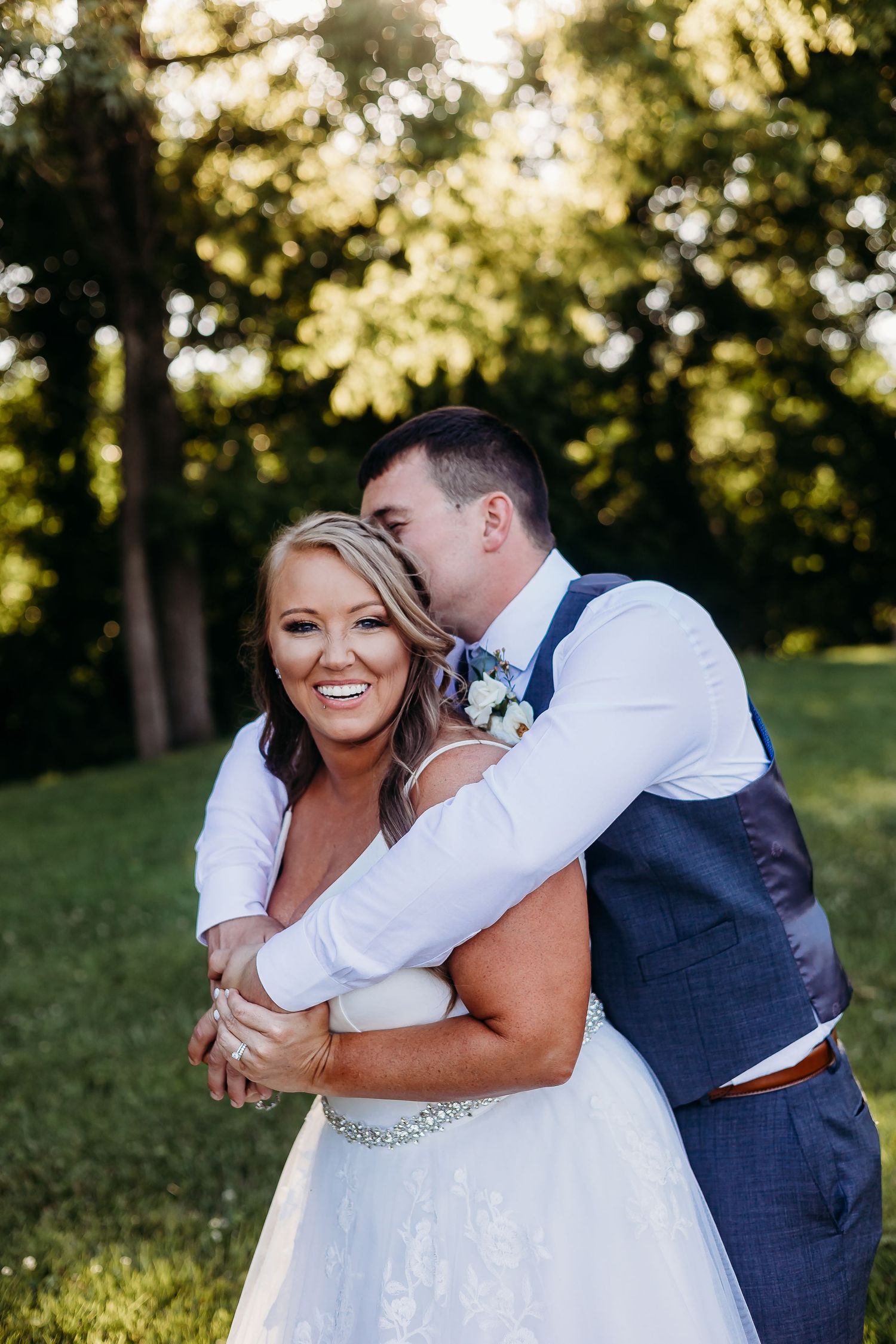 Joyful wedding couple embracing and laughing together in a sunlit outdoor setting.