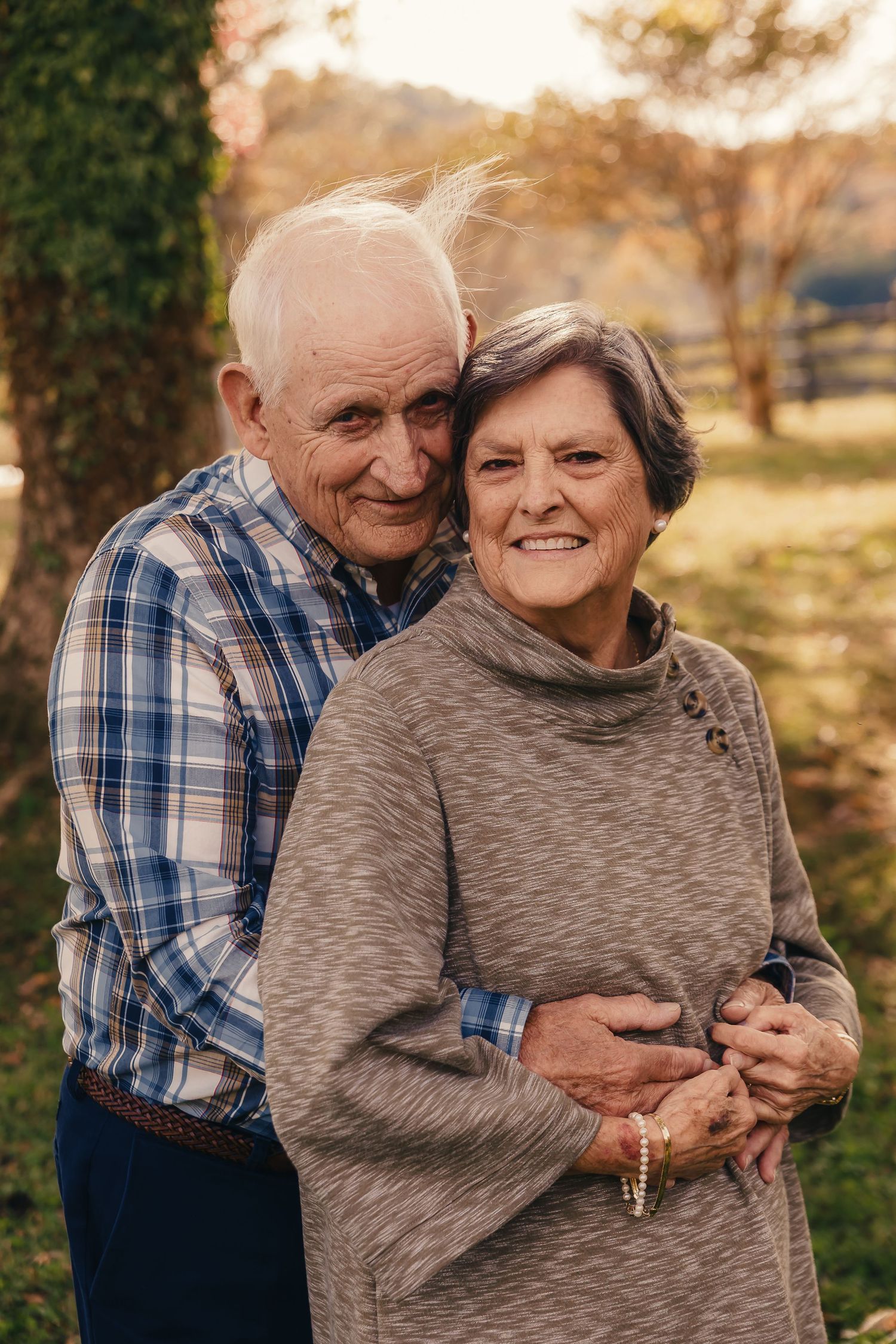 A senior couple embraces while smiling at the camera in an outdoor autumn setting.
