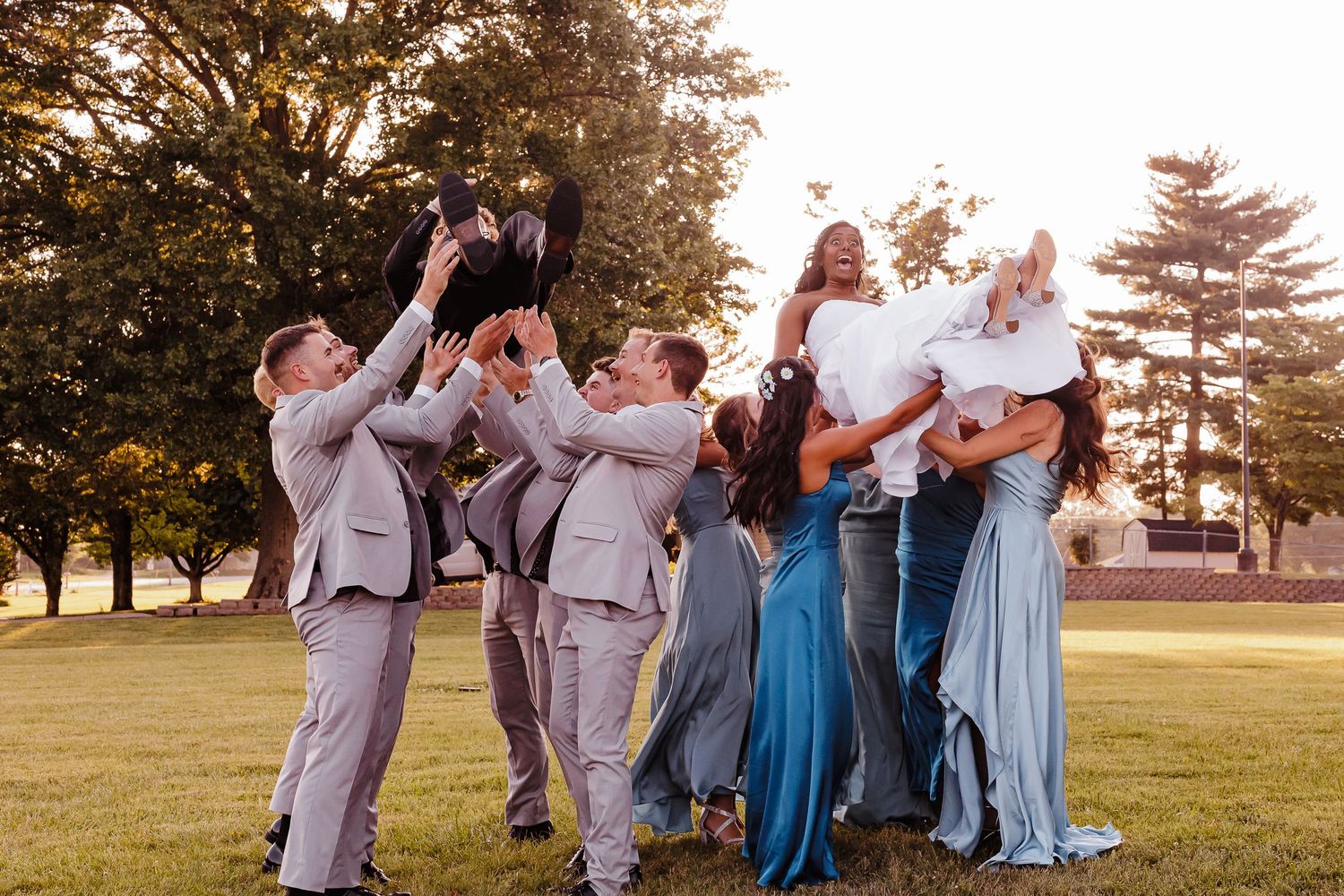 Wedding party celebrates with energetic poses on a grassy lawn at sunset.