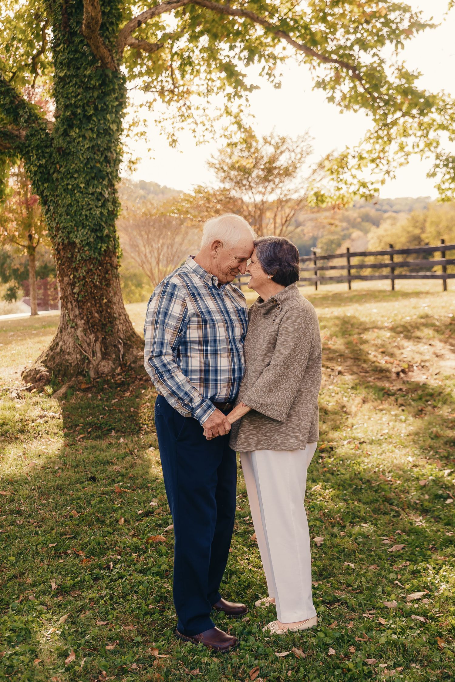 An elderly couple shares an intimate moment under a tree with a rustic fence in the background.