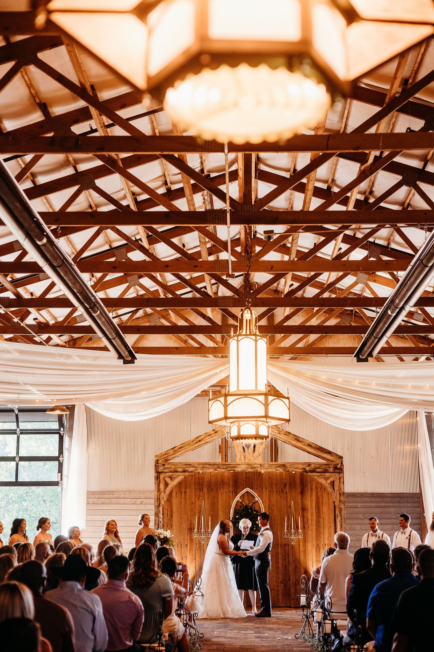 Rustic barn wedding ceremony venue with exposed wooden beams and white fabric draping.