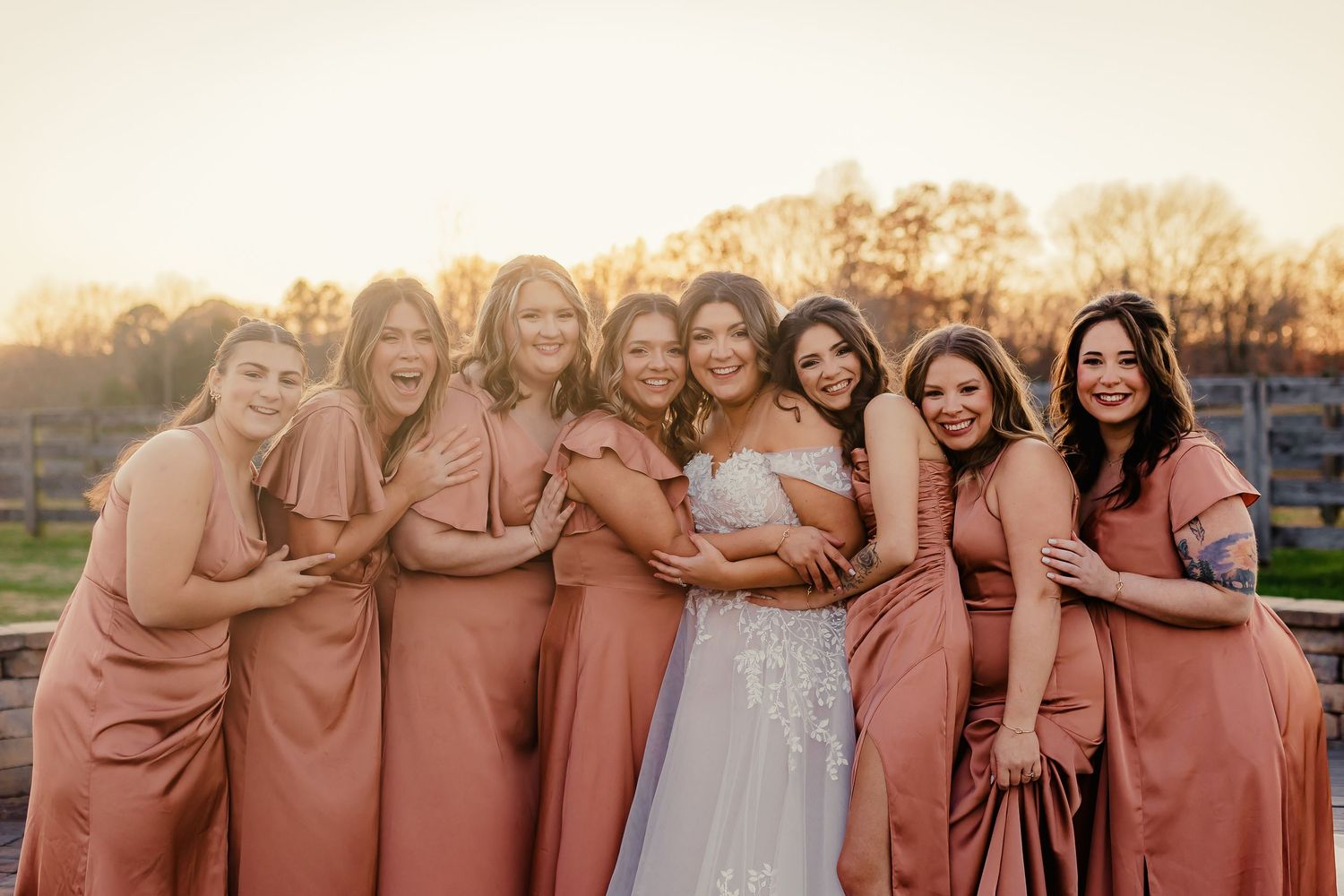 Bridesmaids in dusty rose dresses gather around bride for outdoor group photo.