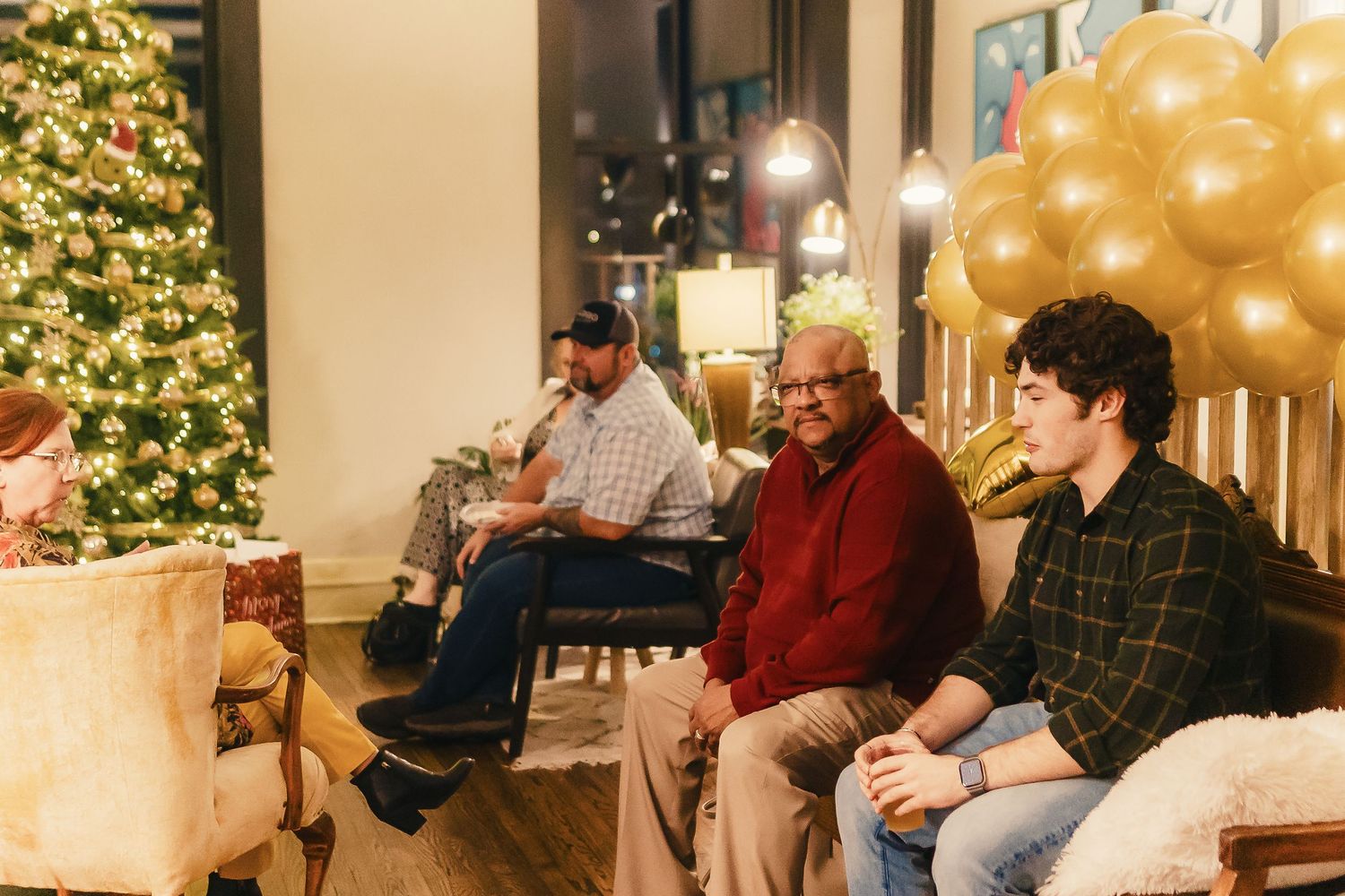 People sit together in a living room decorated for the holidays with a Christmas tree and gold balloons.