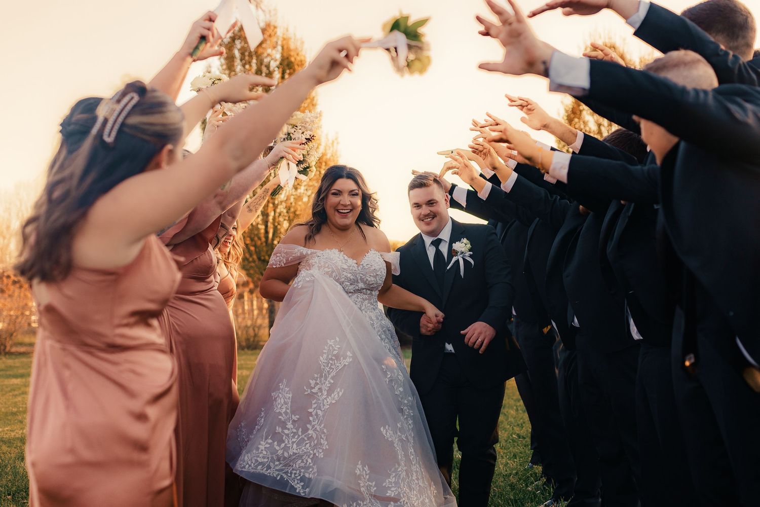 Wedding party celebrates outdoors with raised hands at sunset.