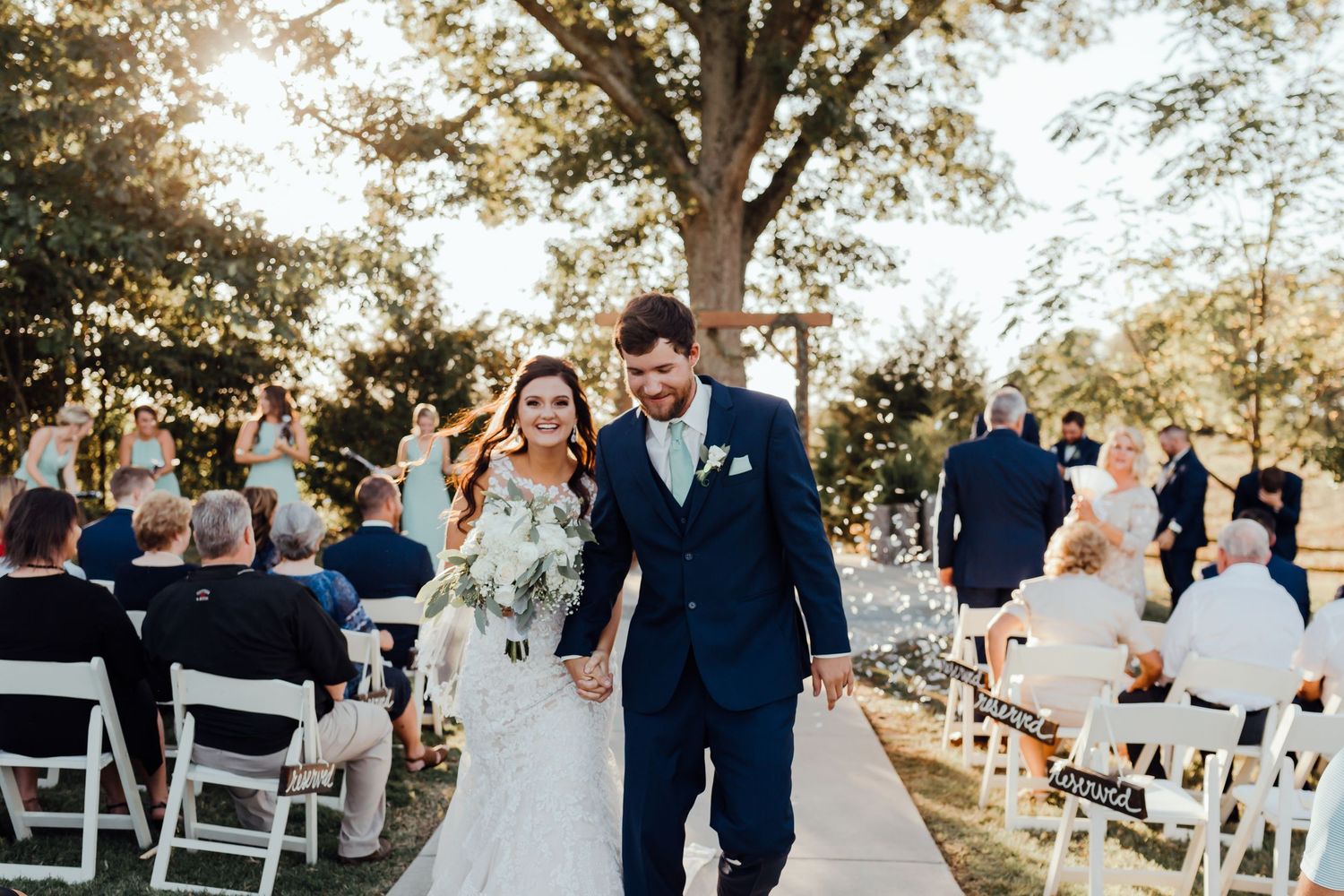 Outdoor wedding ceremony setup with white chairs and natural tree backdrop.