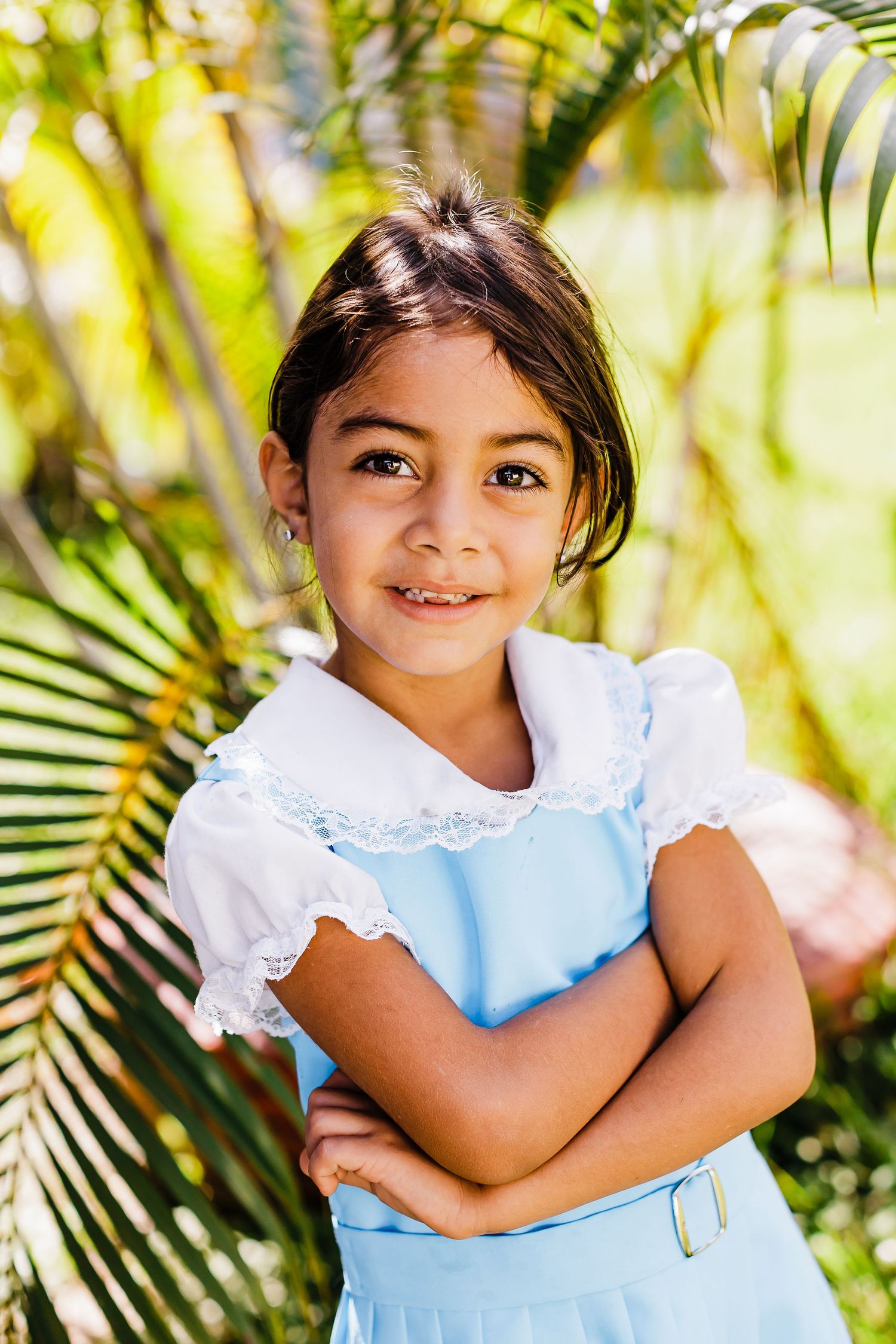 Young girl in a light blue dress smiling against tropical foliage background.