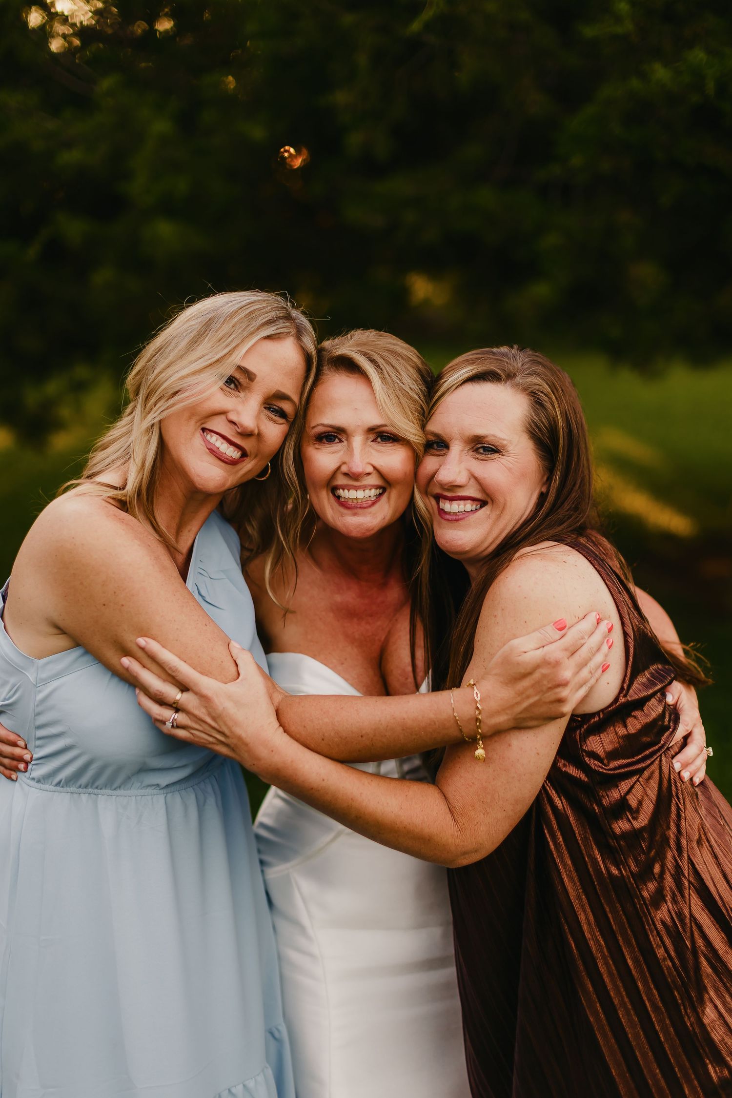 Bride in white gown hugging two smiling women in blue and brown dresses outdoors at golden hour.