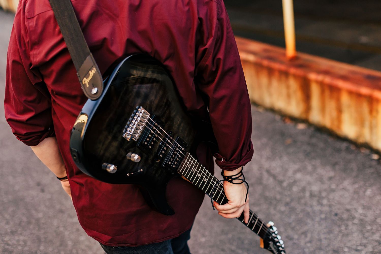 A musician carries a guitar in a black case while wearing a burgundy shirt.
