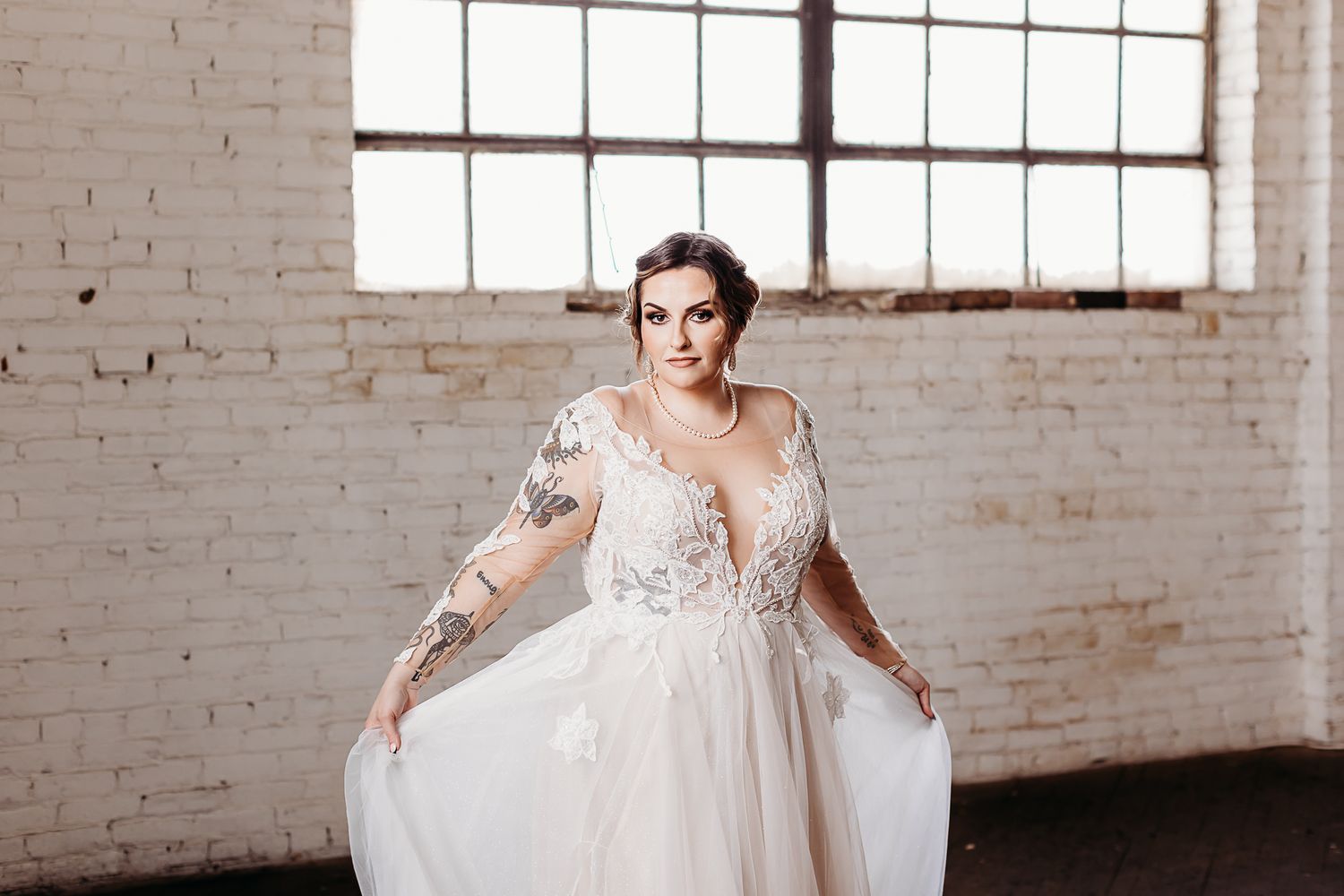 A plus size bride in a lace wedding dress poses in an industrial loft space with exposed brick walls.