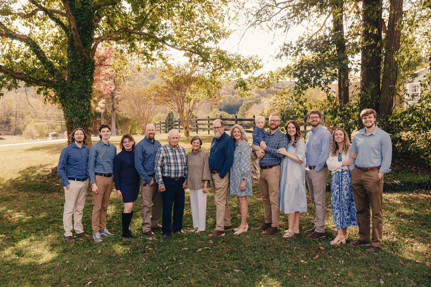 A large family group poses together for a portrait on a sunny autumn day with trees and fencing in the background.