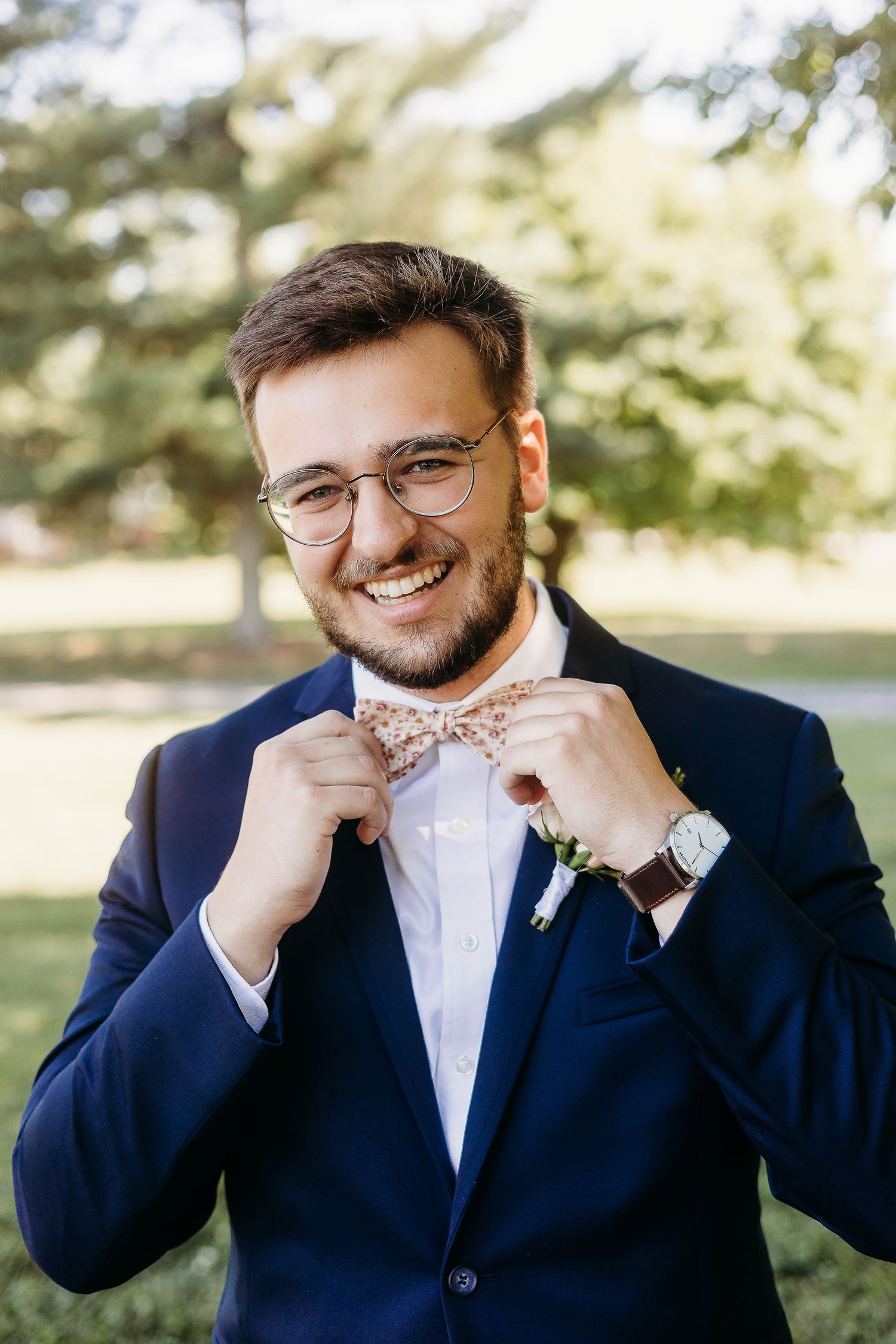 Groom adjusts bowtie while wearing navy suit and glasses outdoors.