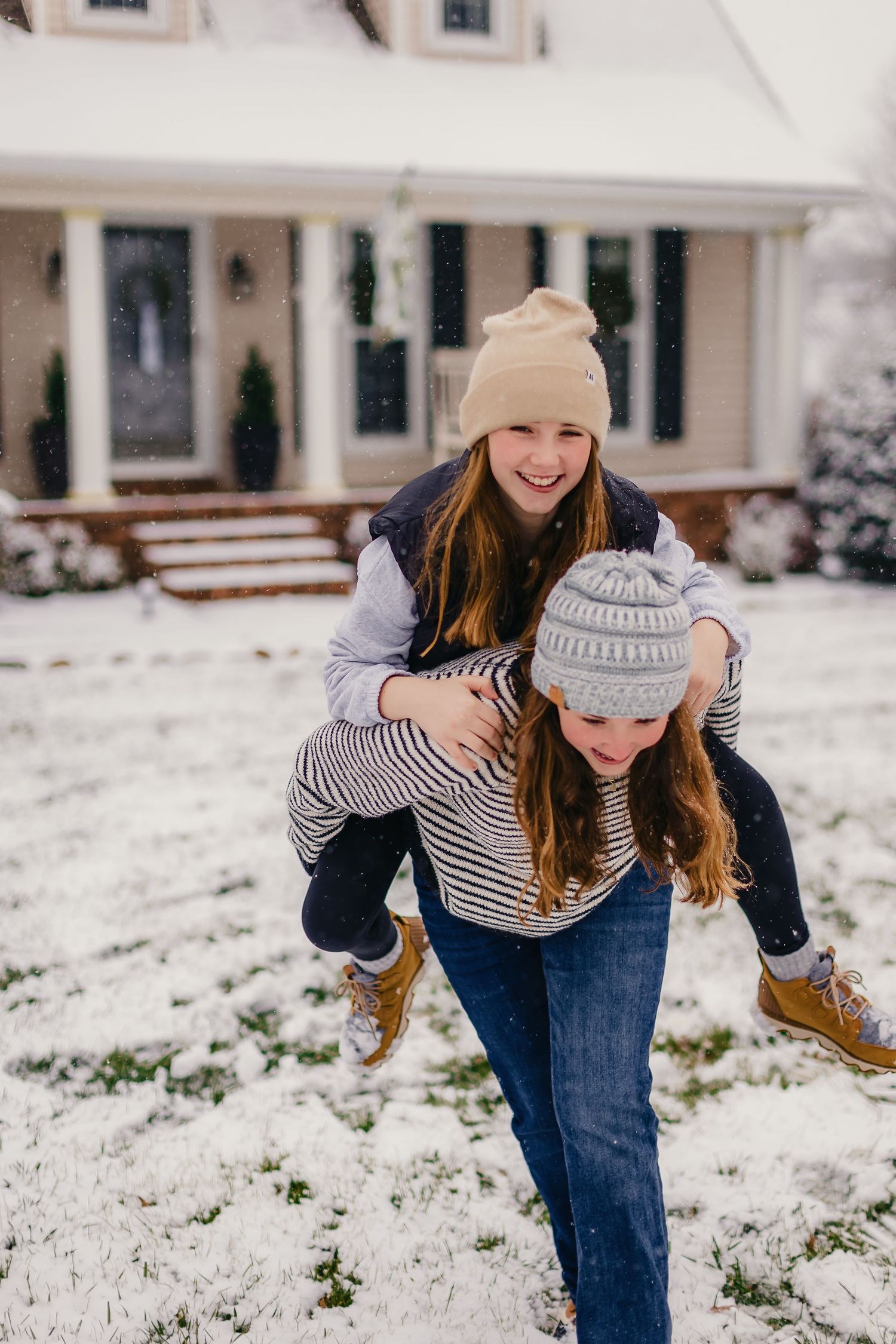 Person in winter clothing bending down playfully in the snow outside a white house.