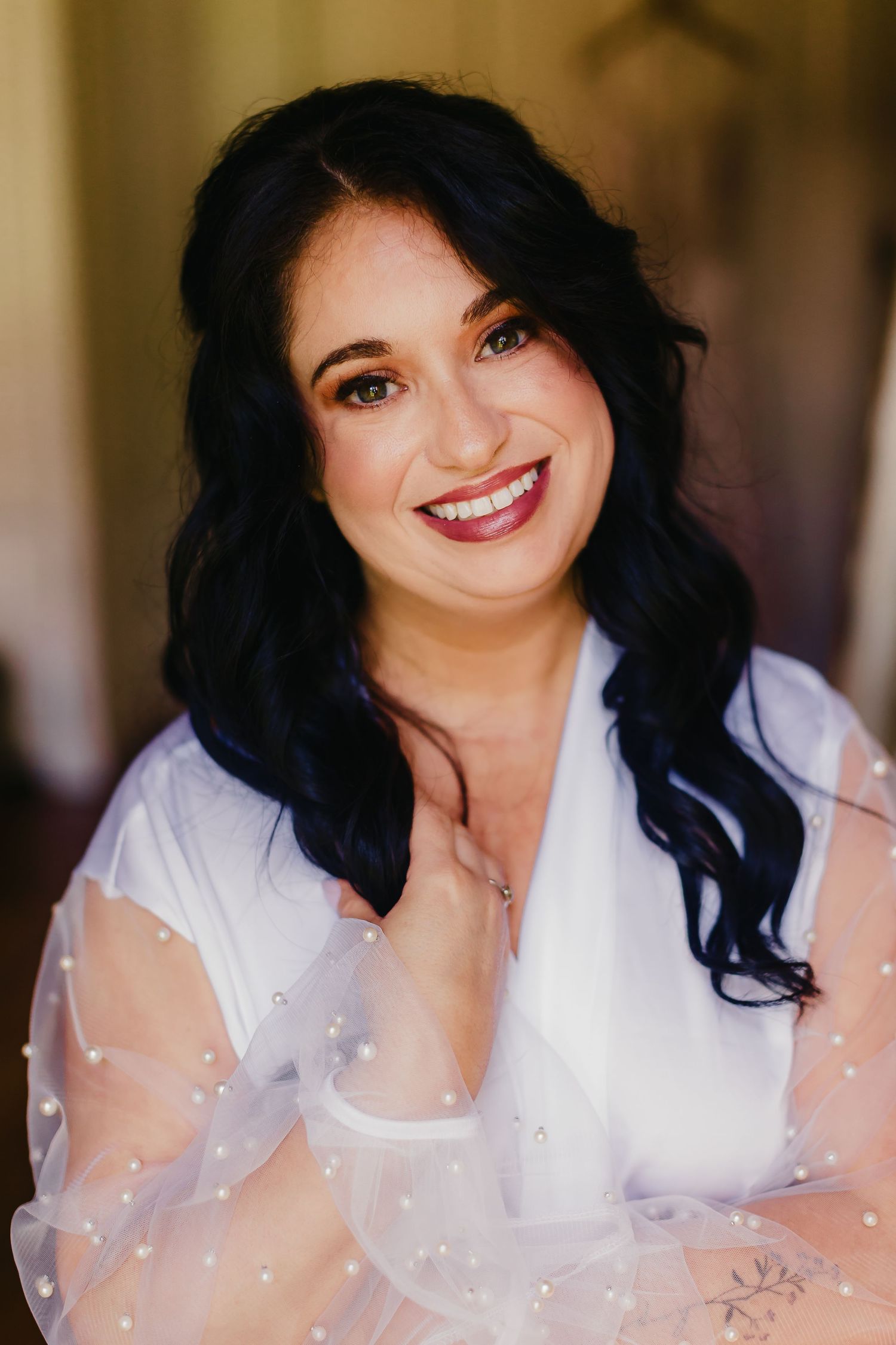 Woman with dark curly hair and red lipstick smiling while wearing a white sheer polka dot blouse.