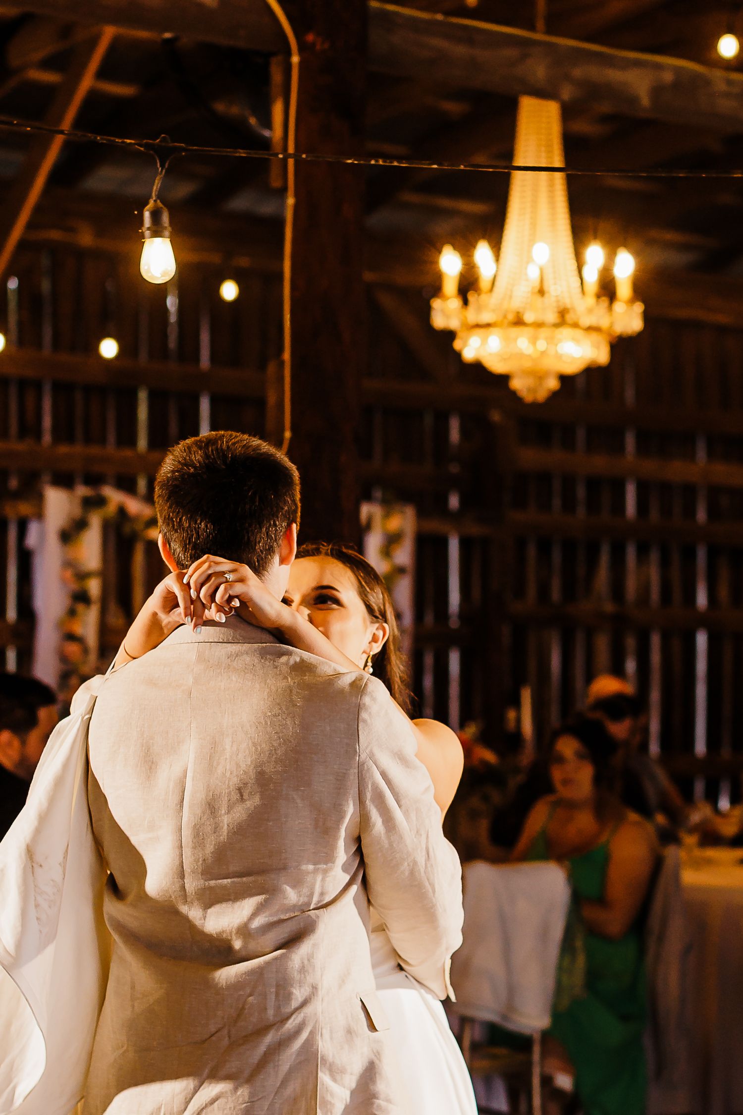 A first dance moment under a crystal chandelier in a rustic barn venue.