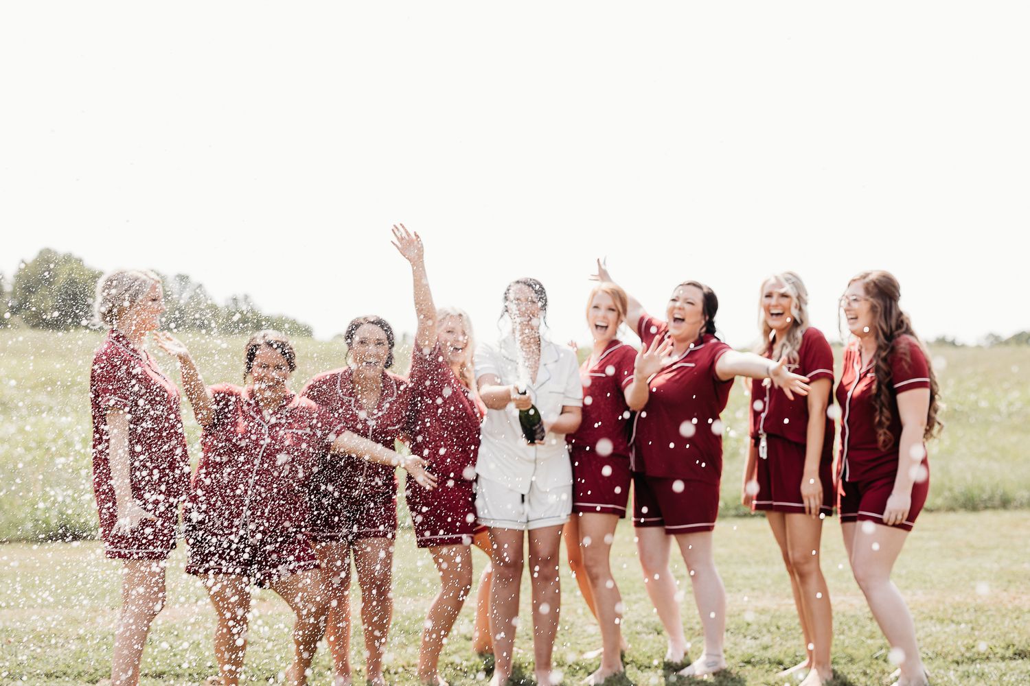 Bridesmaids in burgundy dresses celebrate with champagne spray outdoors.