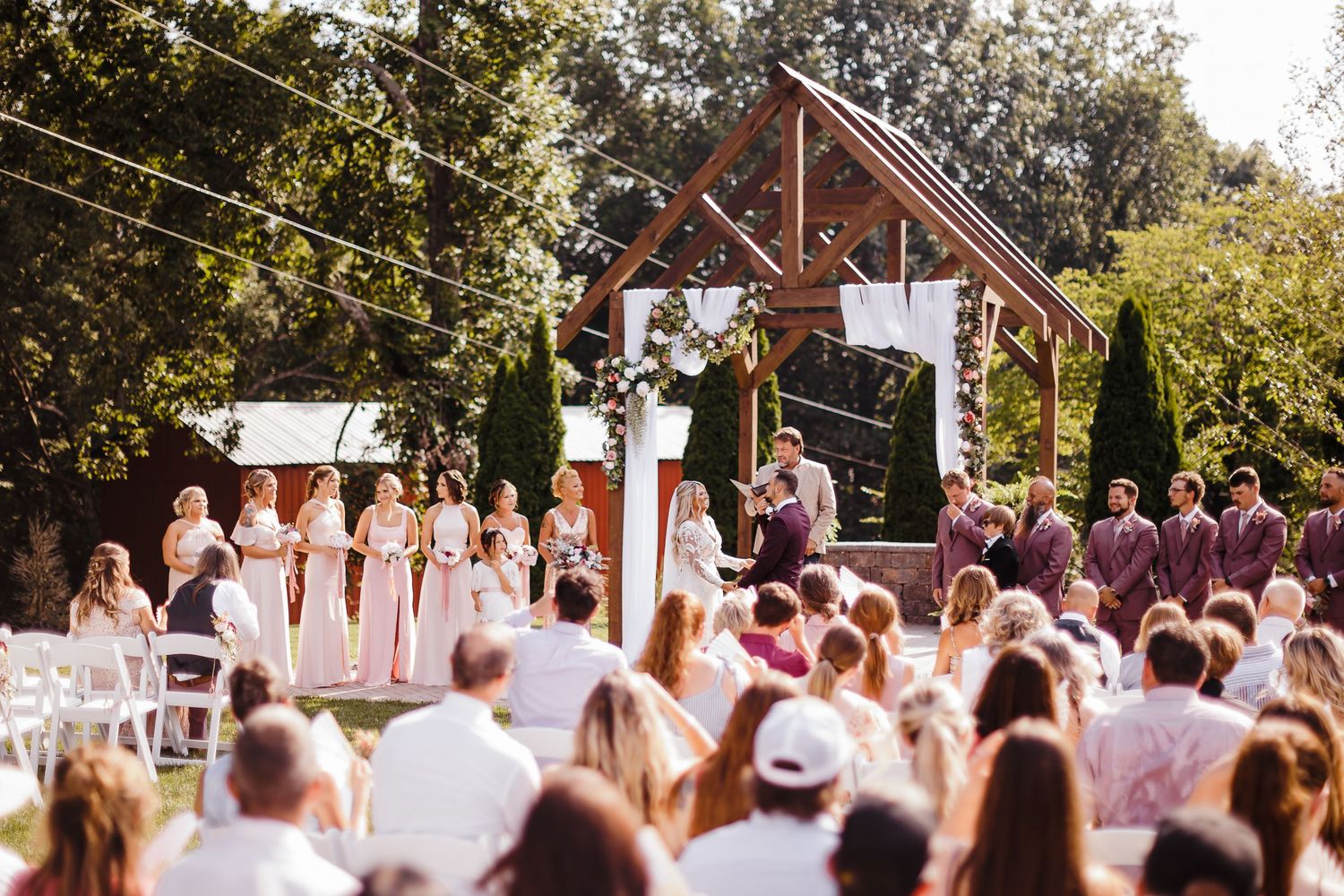 Outdoor wedding ceremony with bridesmaids in pink, groomsmen in burgundy, under a wooden arch surrounded by lush greenery.