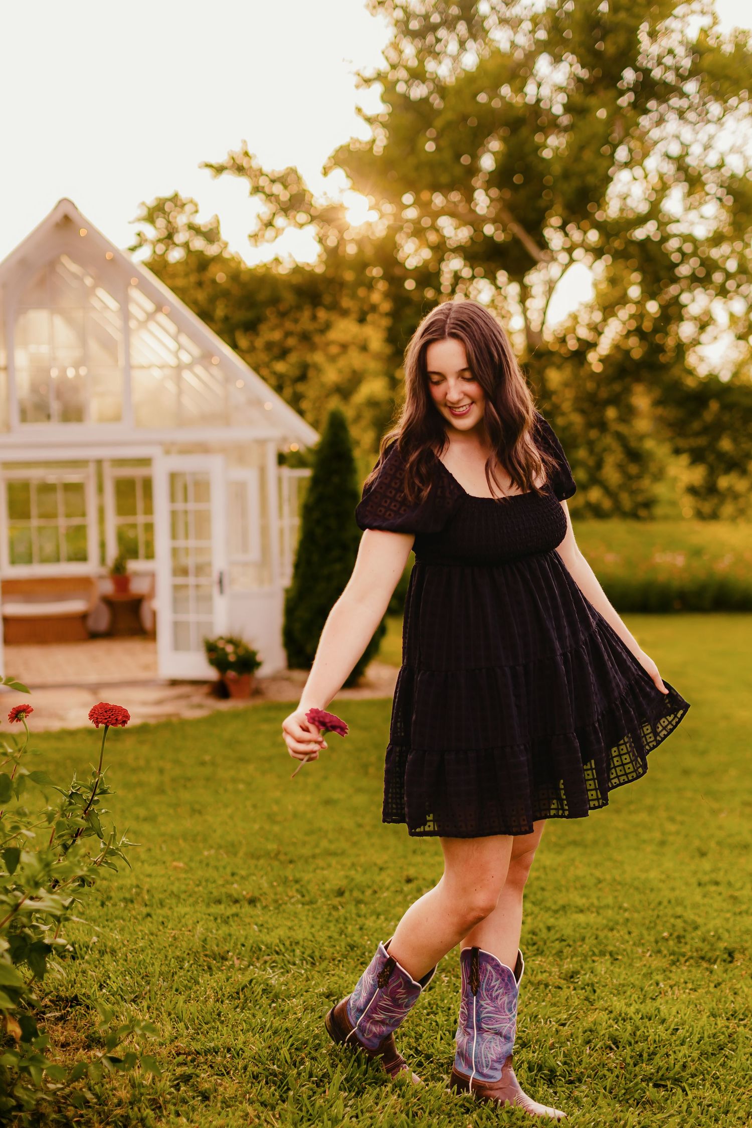 Young woman in black dress and cowboy boots holding a flower in a sunny garden with greenhouse backdrop.