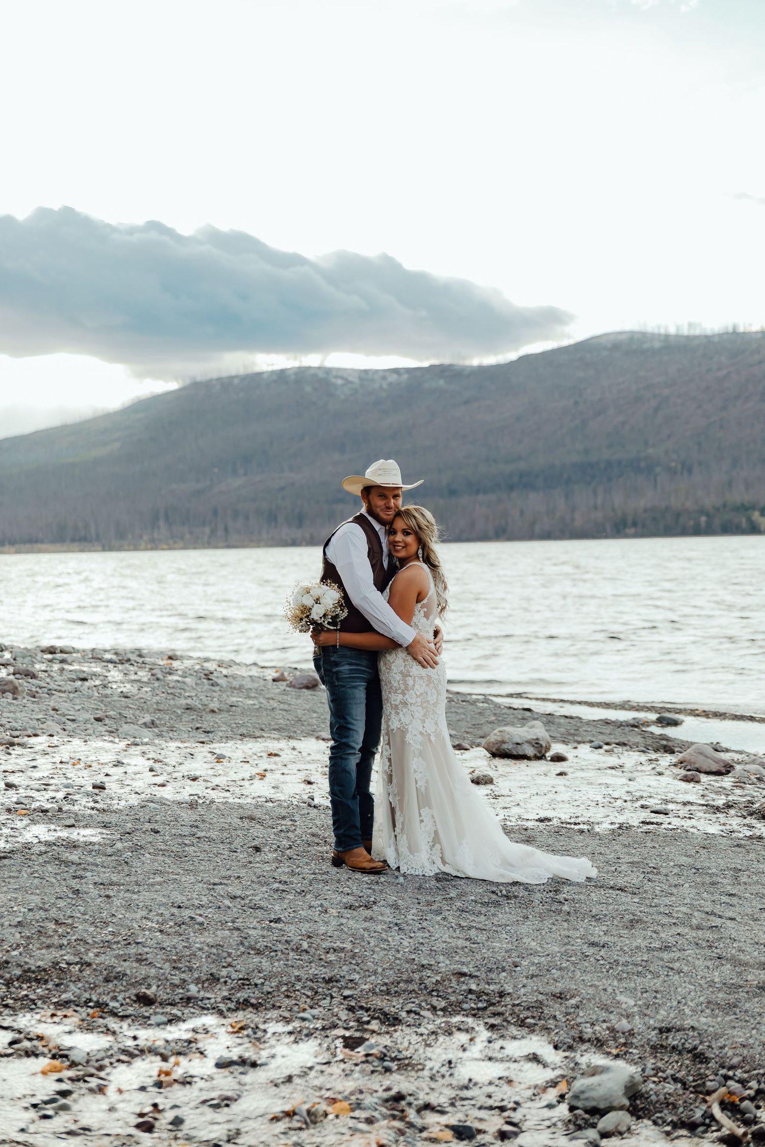 A couple shares an intimate moment on a rocky beach with mountains and cloudy skies in the background.
