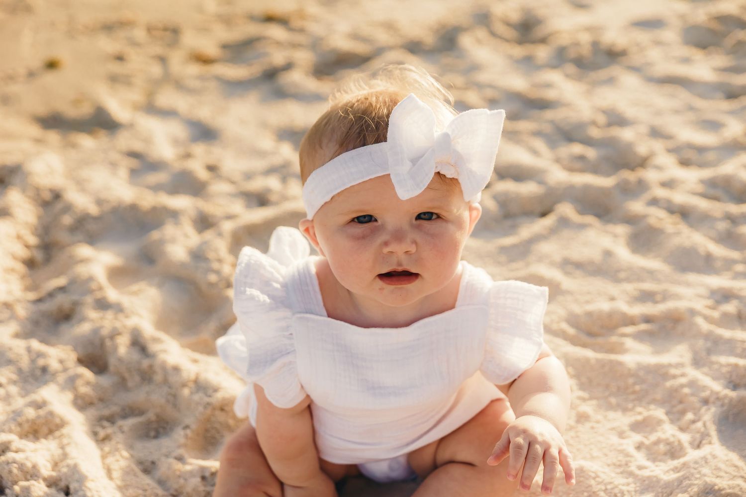 Baby in white outfit sits on sandy beach during golden hour portrait session.