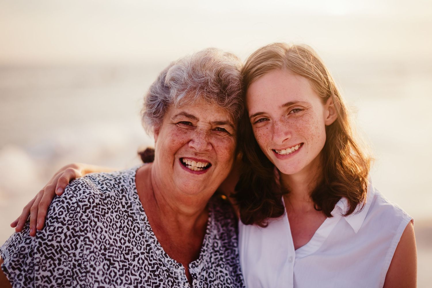 Two women smiling together on a beach at sunset, an older woman with gray hair and a younger redhead.