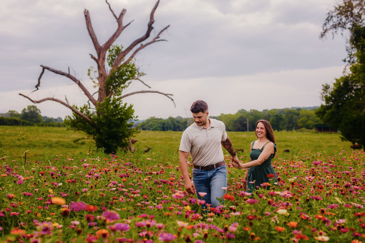 Couple walking hand in hand through a vibrant wildflower meadow with a dead tree backdrop on a cloudy day.