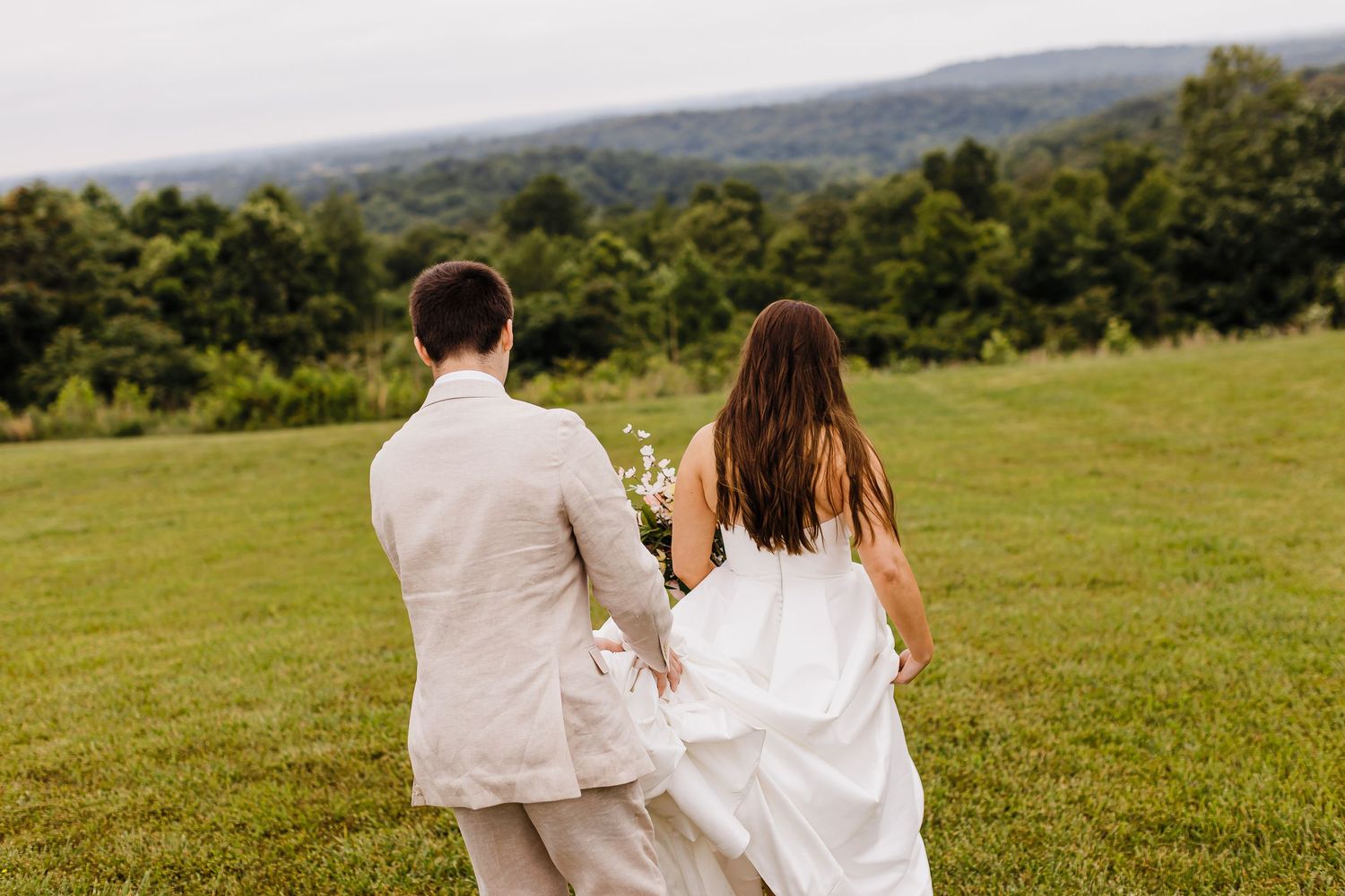 A couple in wedding attire walks through a scenic hilltop meadow.