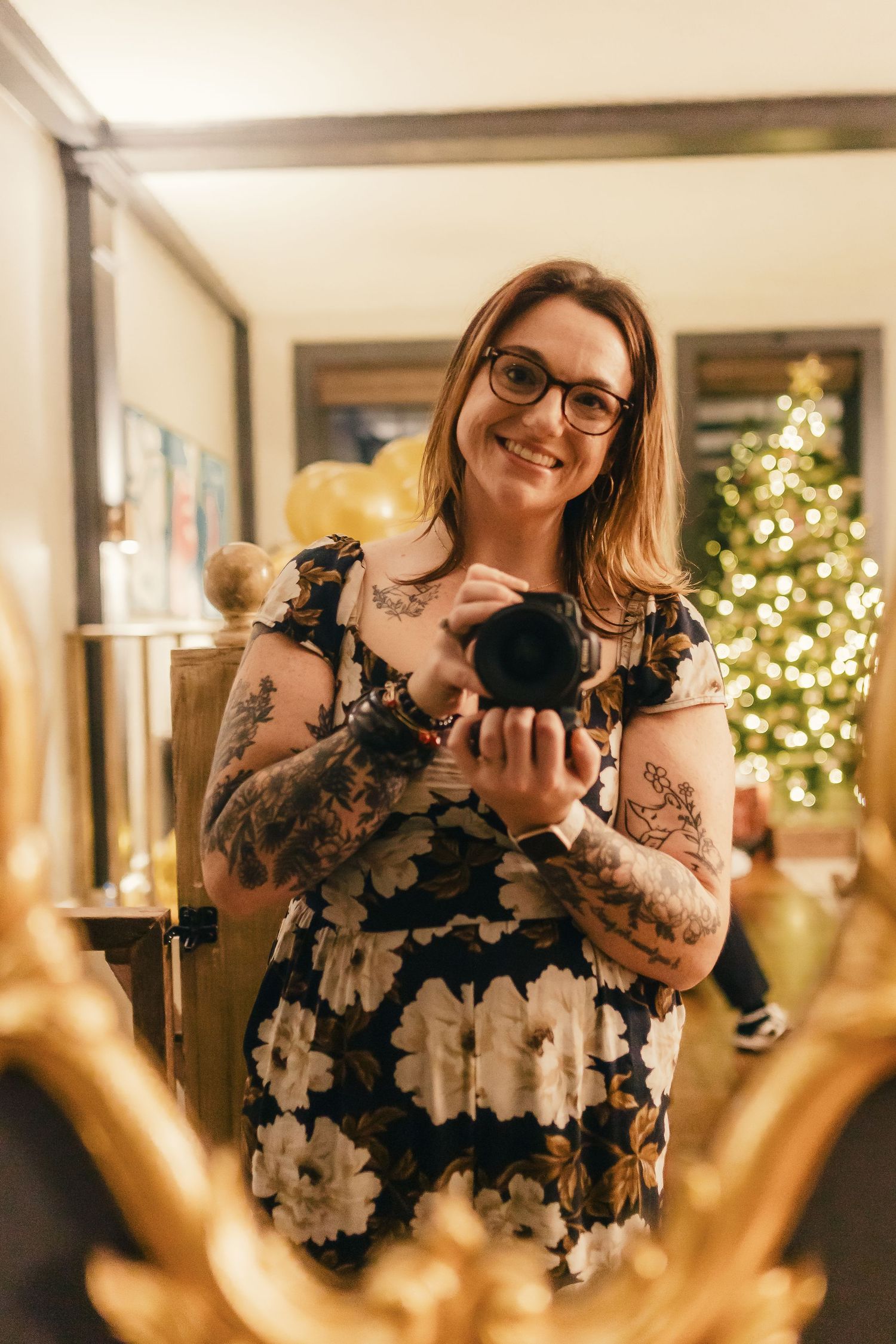A person in a floral dress with tattoos holds a camera while standing in front of a Christmas tree.
