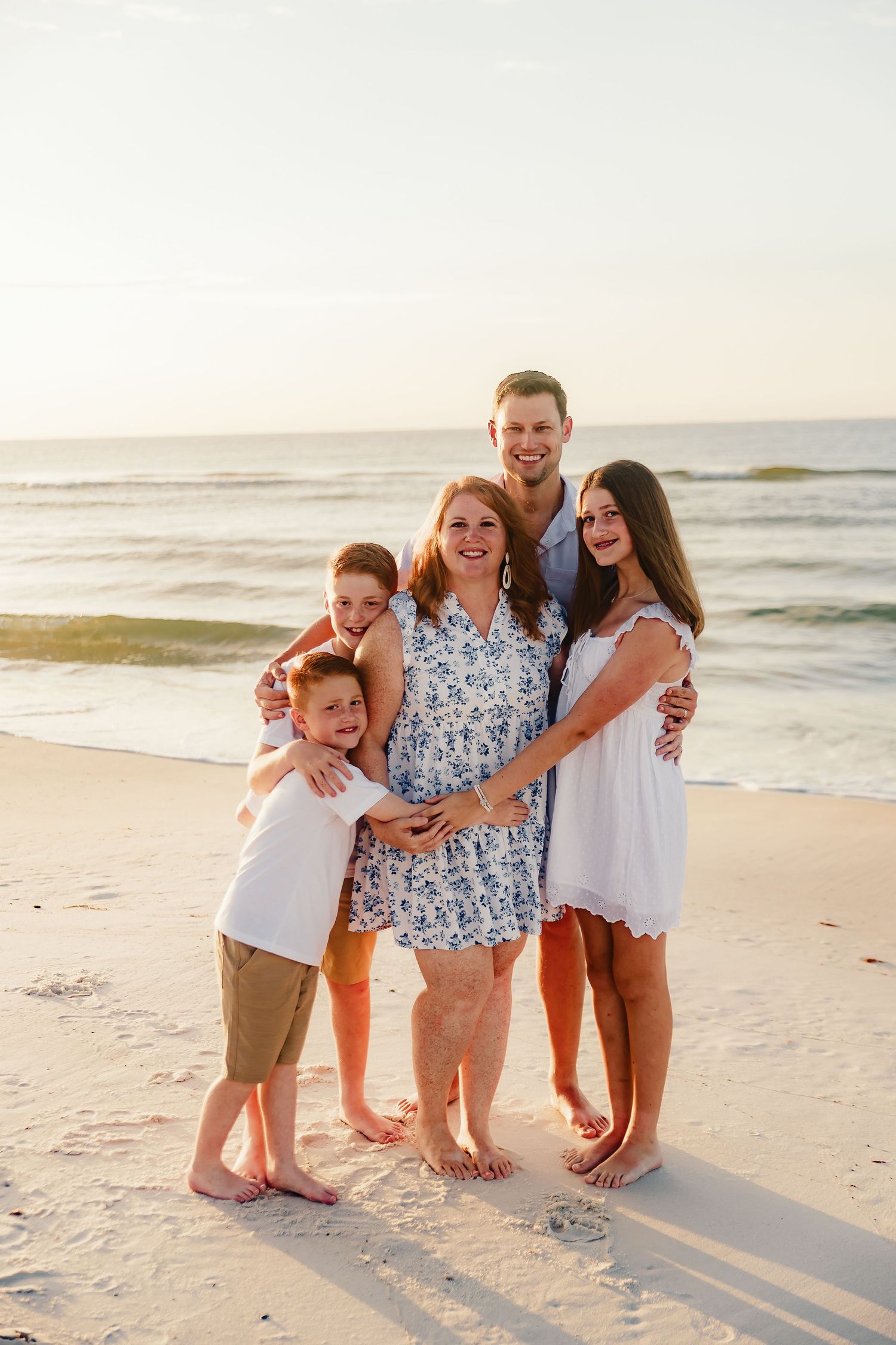 Family group poses together on sandy beach at sunset wearing coordinated white and blue outfits.