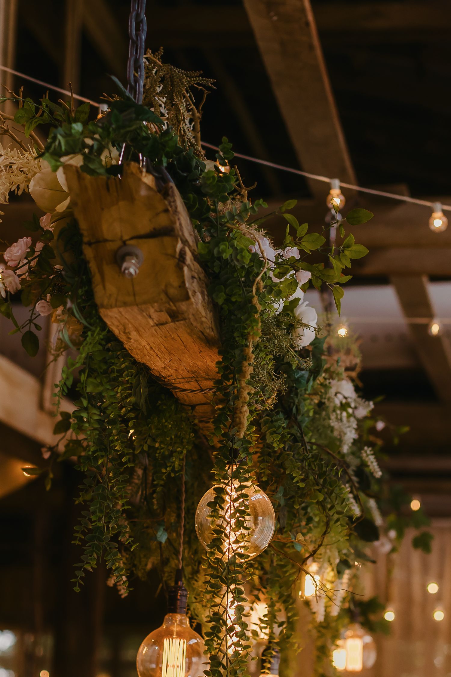 Rustic barn wedding decor featuring hanging greenery, twinkle lights, and vintage-style Edison bulbs creating ambient lighting.