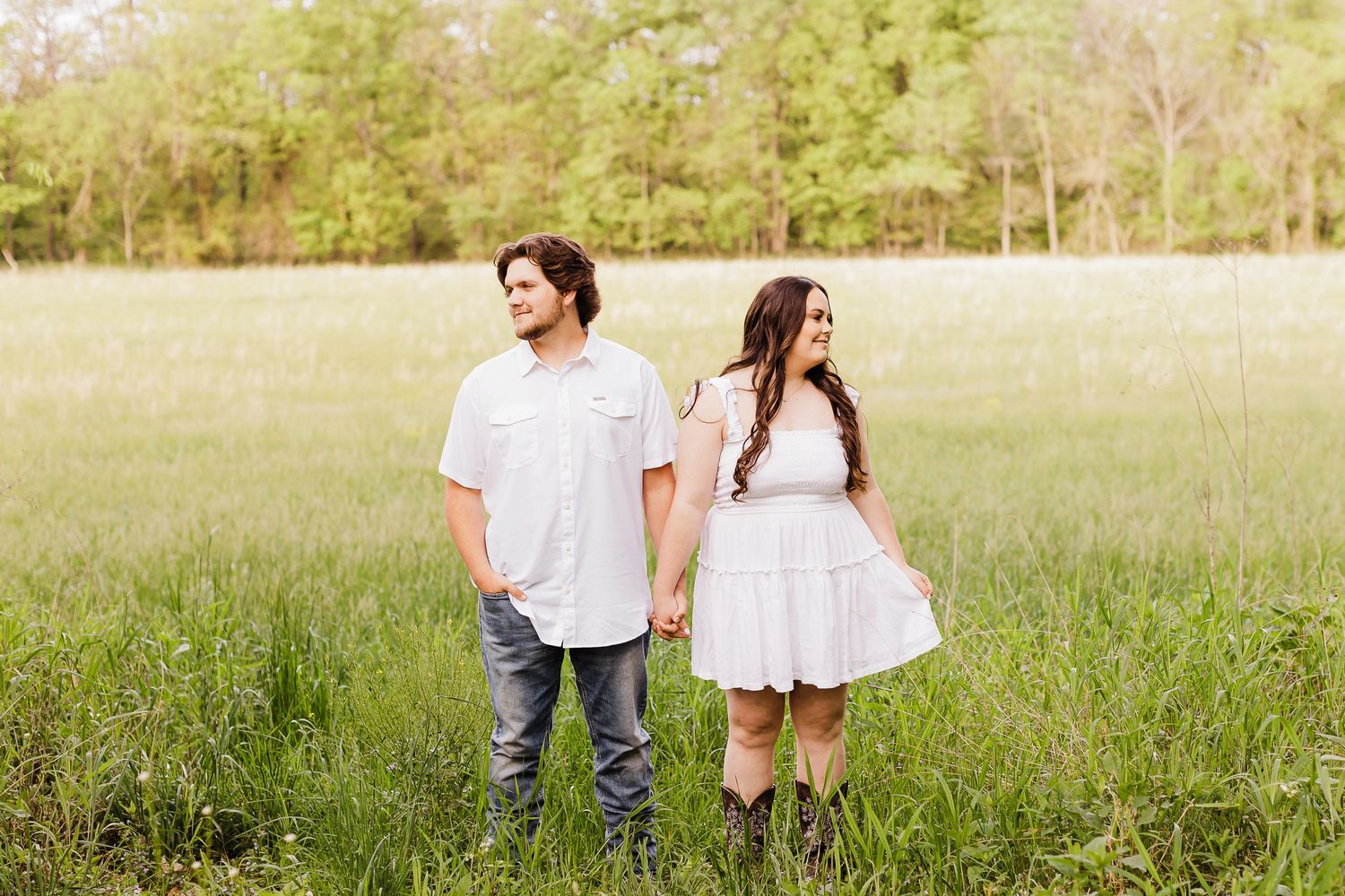 A couple in white clothing stands together in a sunlit grassy field.