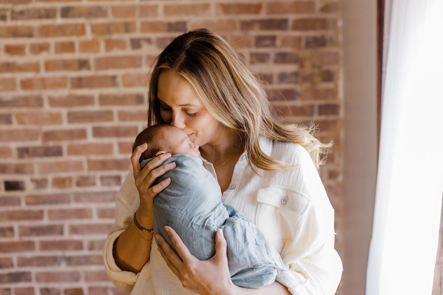 A tender moment captured as a parent in white embraces and kisses a baby in soft blue against a brick wall backdrop.